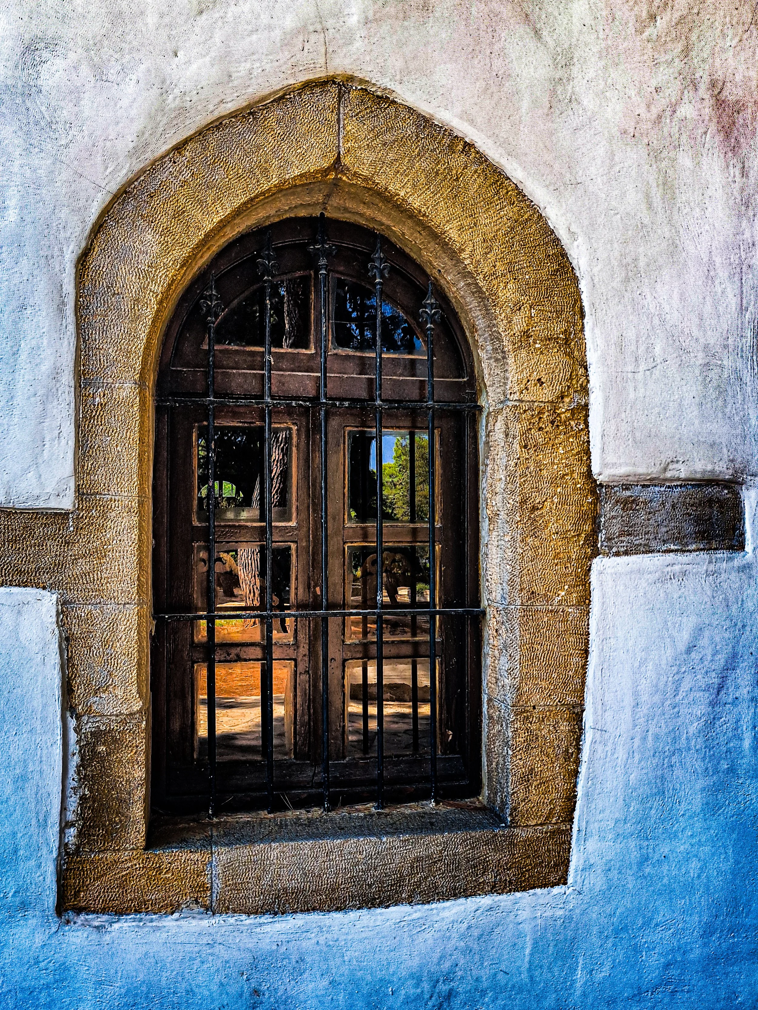 Close-up of a small, arched, wooden window with black metal bars, set into a textured stone wall with a white plaster finish.