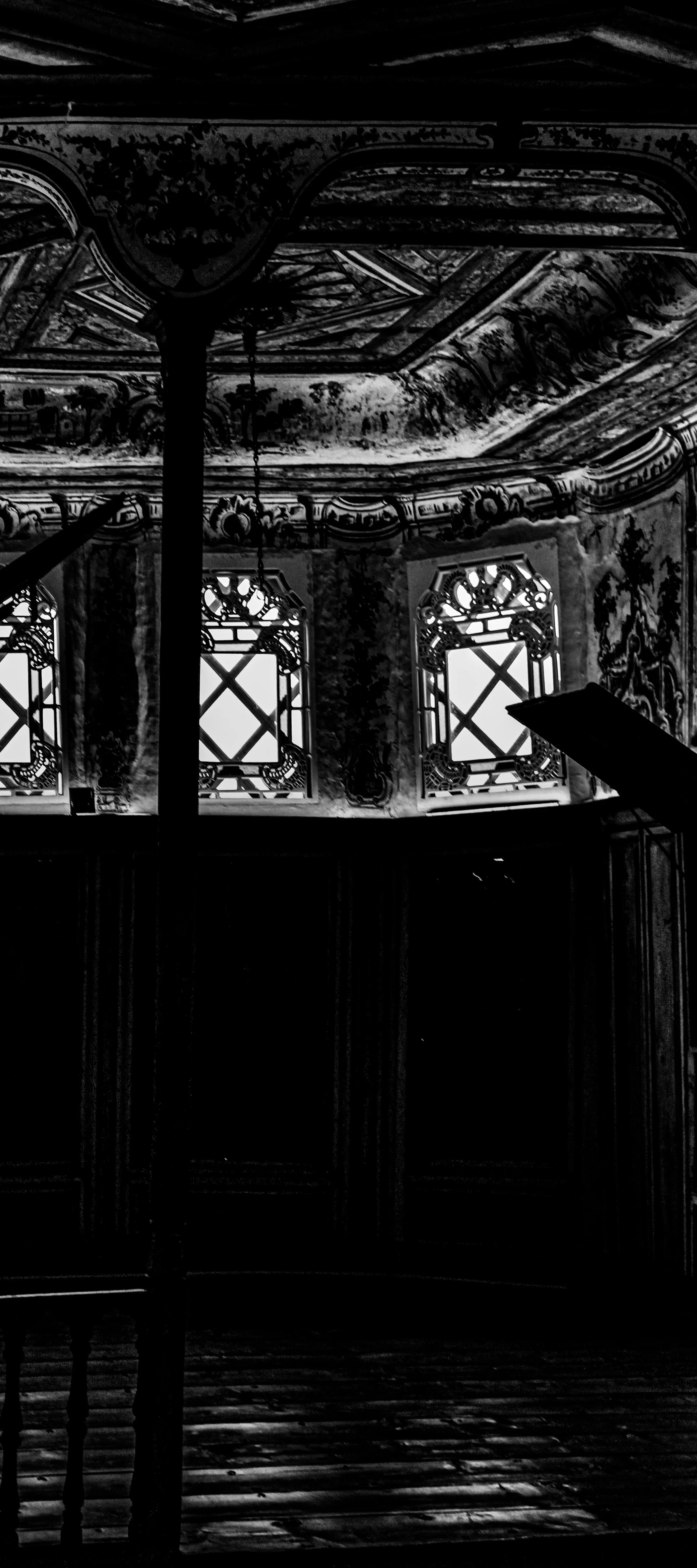 Black and white photograph of an ornate, decorated room interior with detailed ceiling and decorative windows. The room appears dimly lit, with light coming through the windows, casting shadows on the wooden floor.