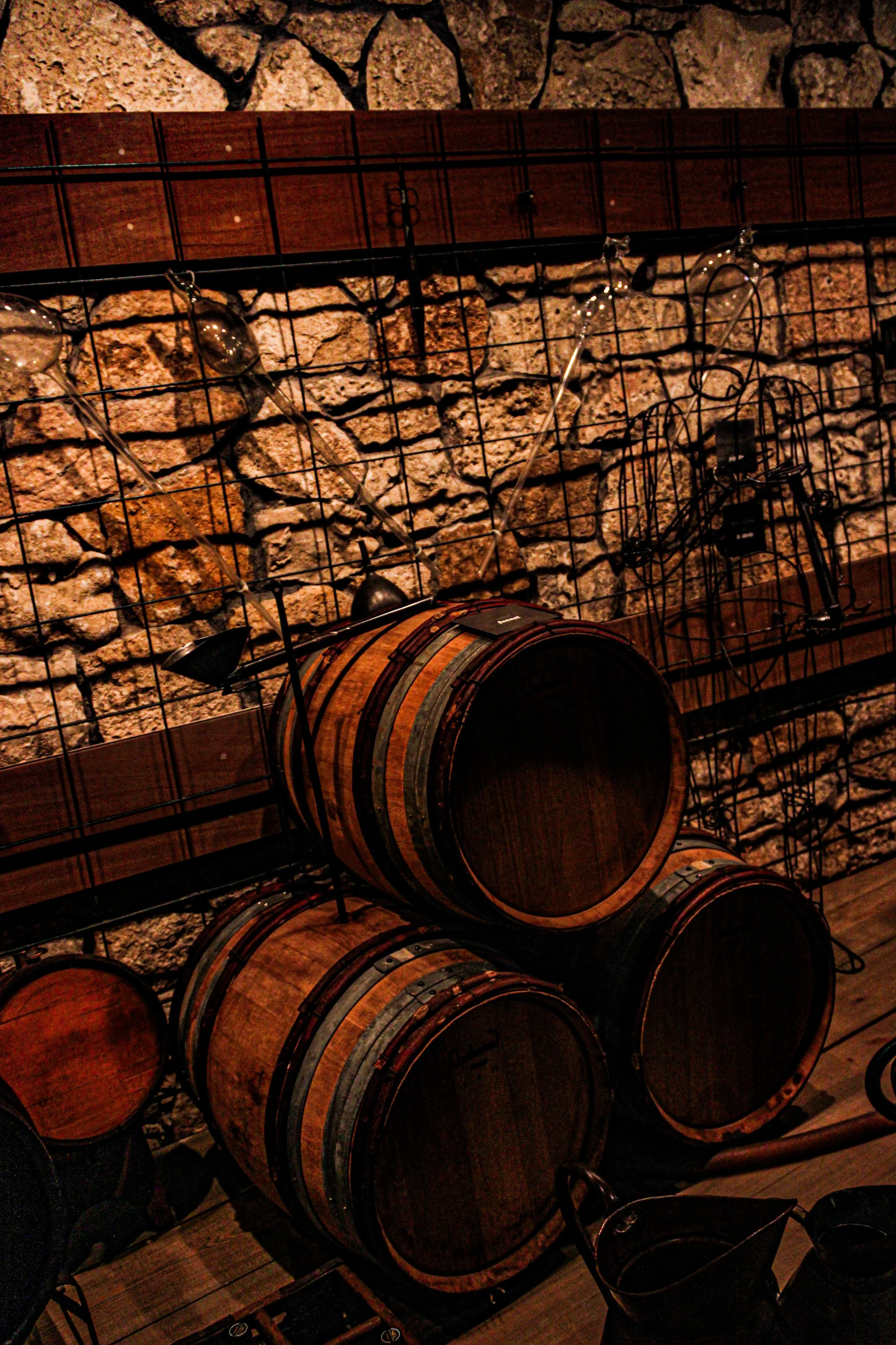 A rustic indoor scene with a stone wall, metal wine racks filled with wooden barrels, and a few empty wine glasses on a table.