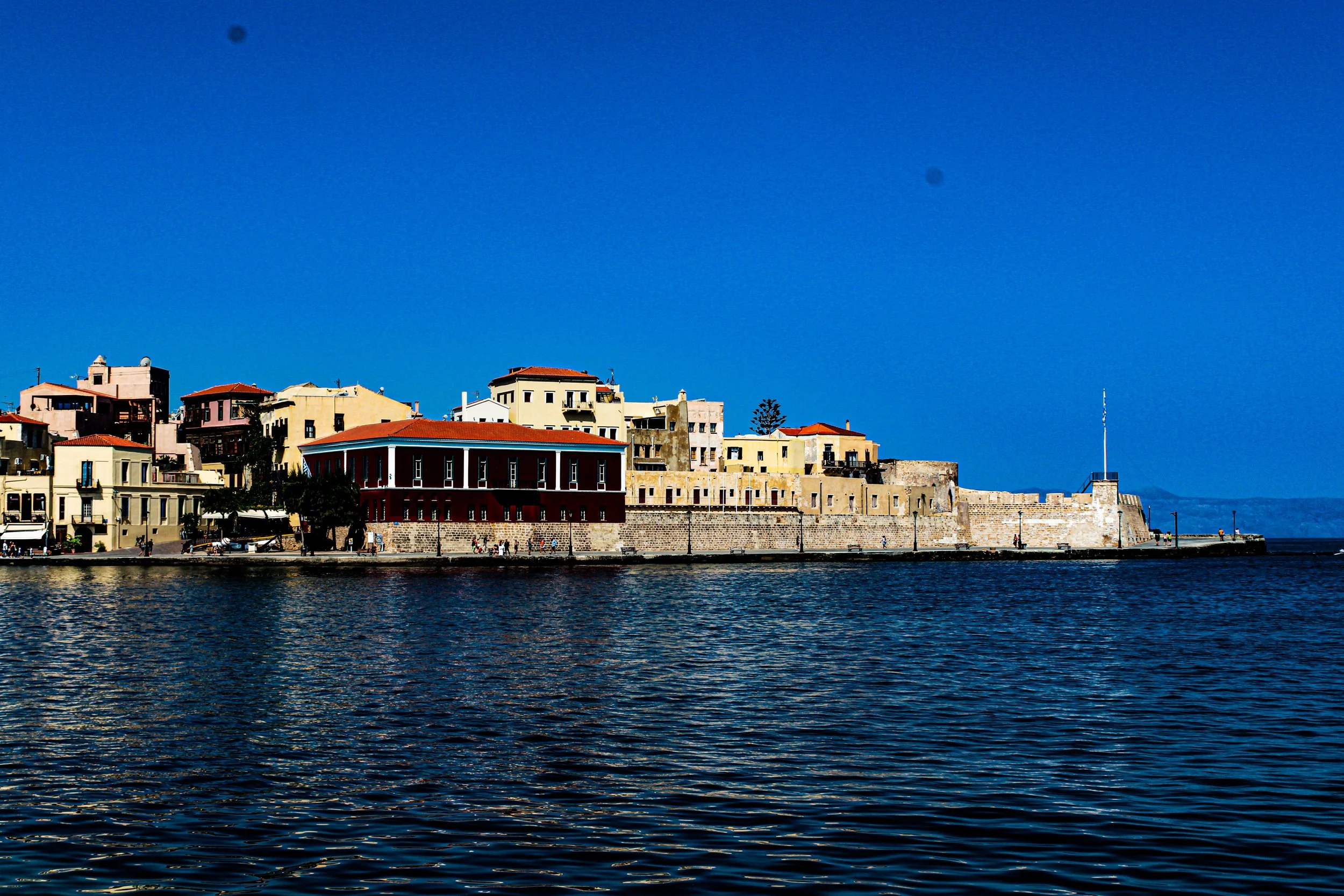 A waterfront view of a city with colorful buildings and a stone wall border, under a clear blue sky.