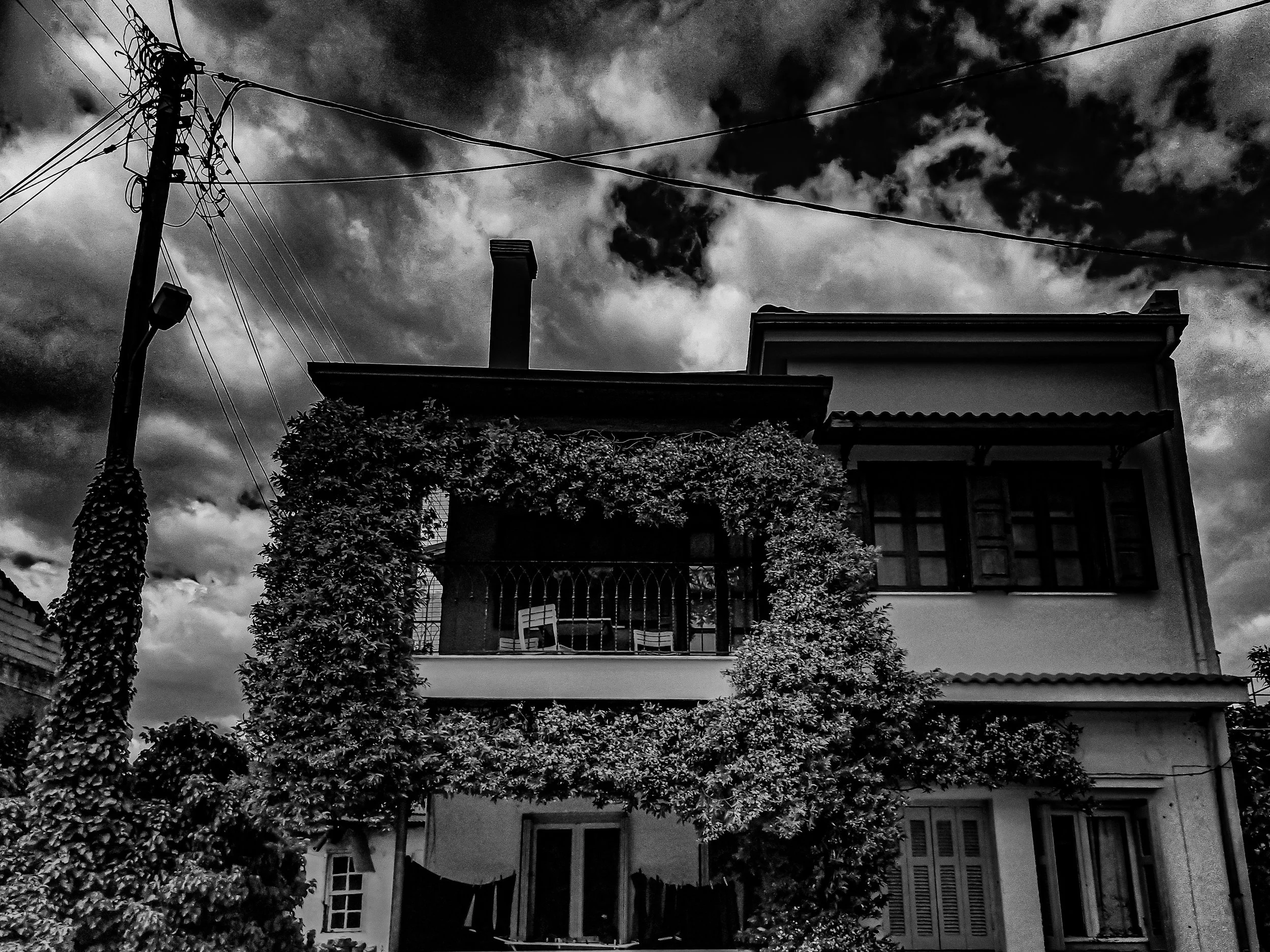 A black and white photo of a house with a plant-covered front porch, a chimney, and a balcony. There is an electrical pole with wires in the foreground and a cloudy sky in the background.