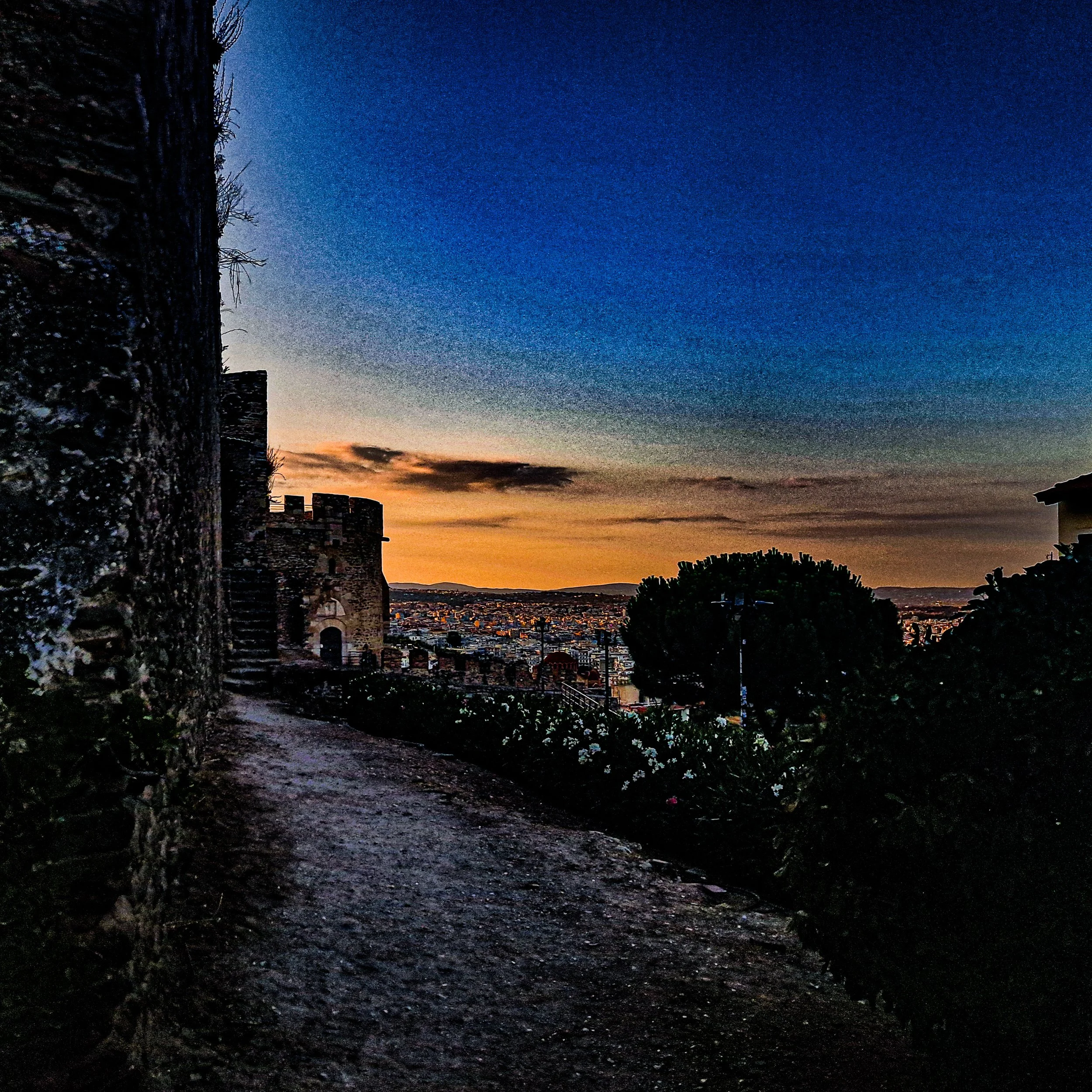 A dirt pathway alongside aged stone walls with plants growing on them, leading to a cityscape at dusk with a sky transitioning from sunset colors to dark blue.