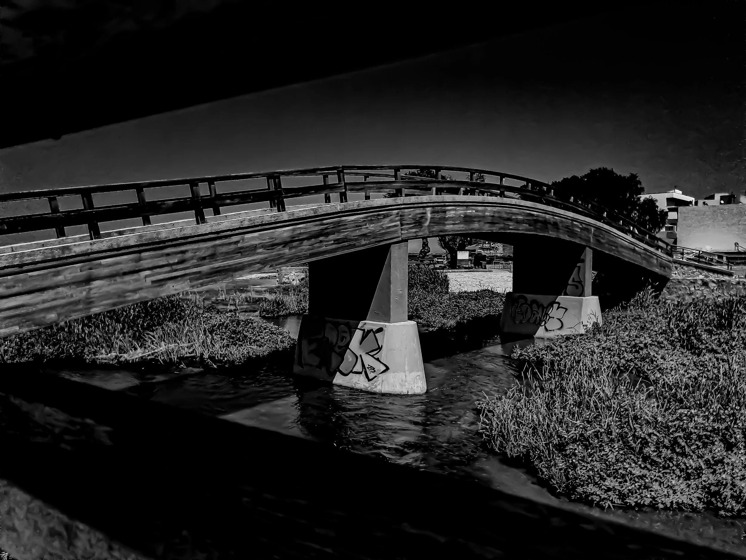 Nighttime scene of a wooden bridge over a small stream with graffiti on the supports, trees, and a building in the background.