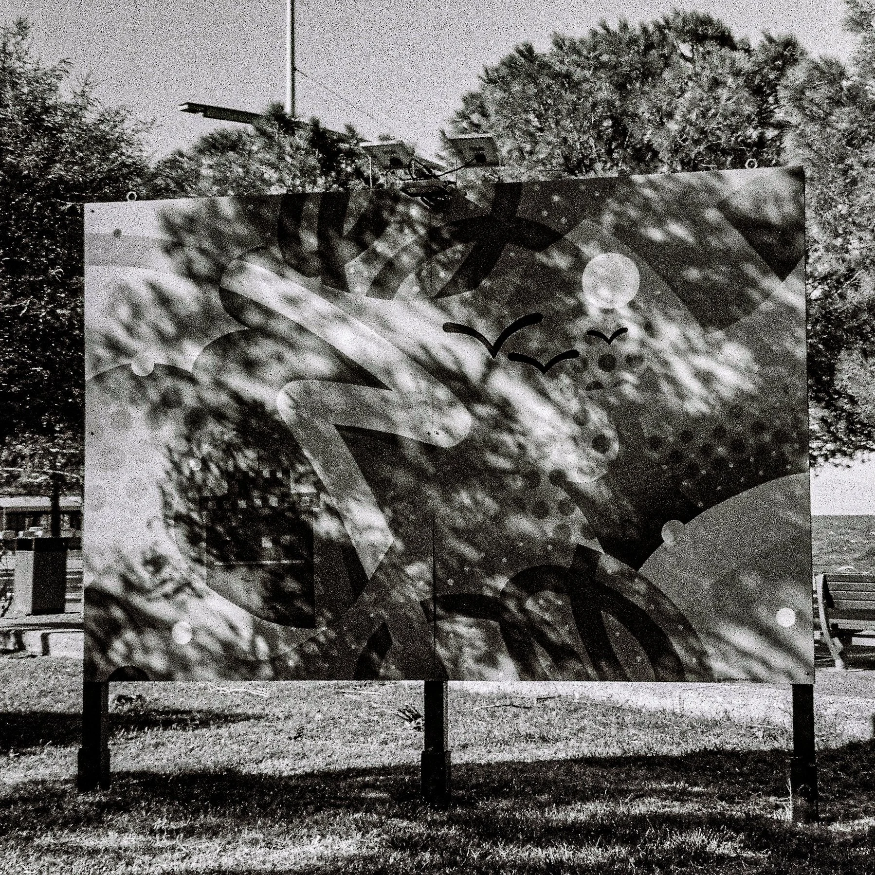 Black and white outdoor mural artwork on a wall, featuring various abstract patterns and shapes, with trees and wires visible in the background.