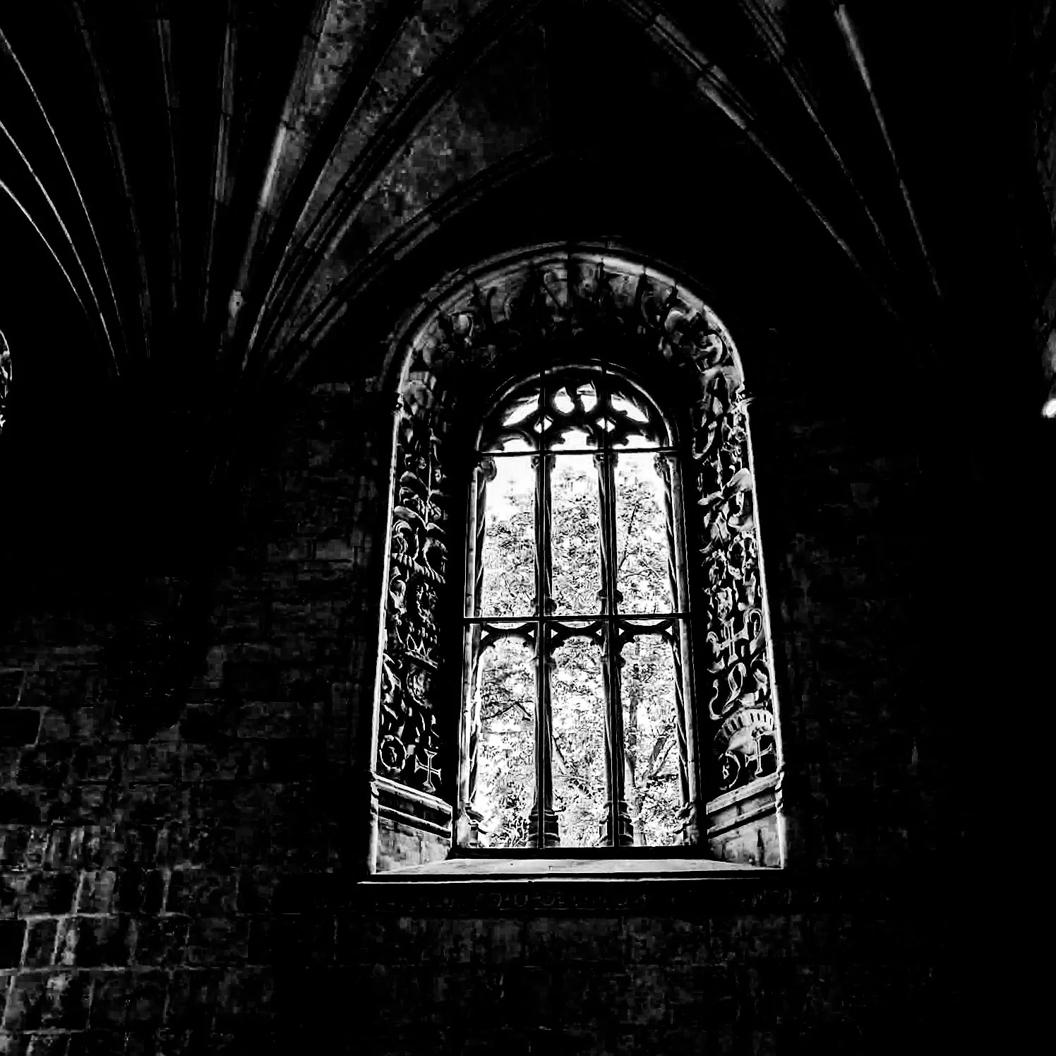 Black-and-white photo of an ornate, Gothic-style window with intricate carvings, set in a dark stone wall, showing trees and sky outside.