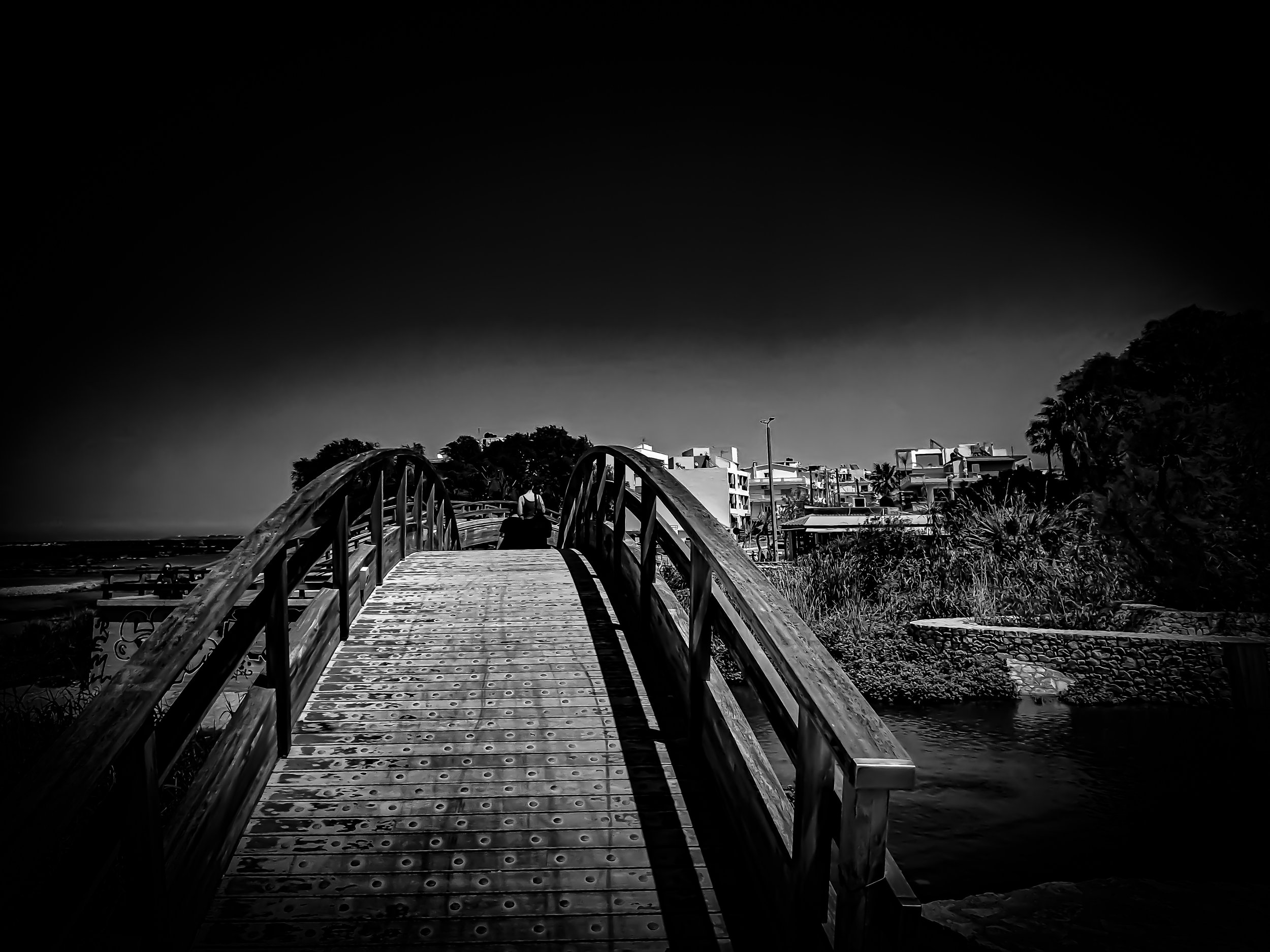 Black and white photo of a person standing on a pier or bridge over water, with buildings and trees in the background.