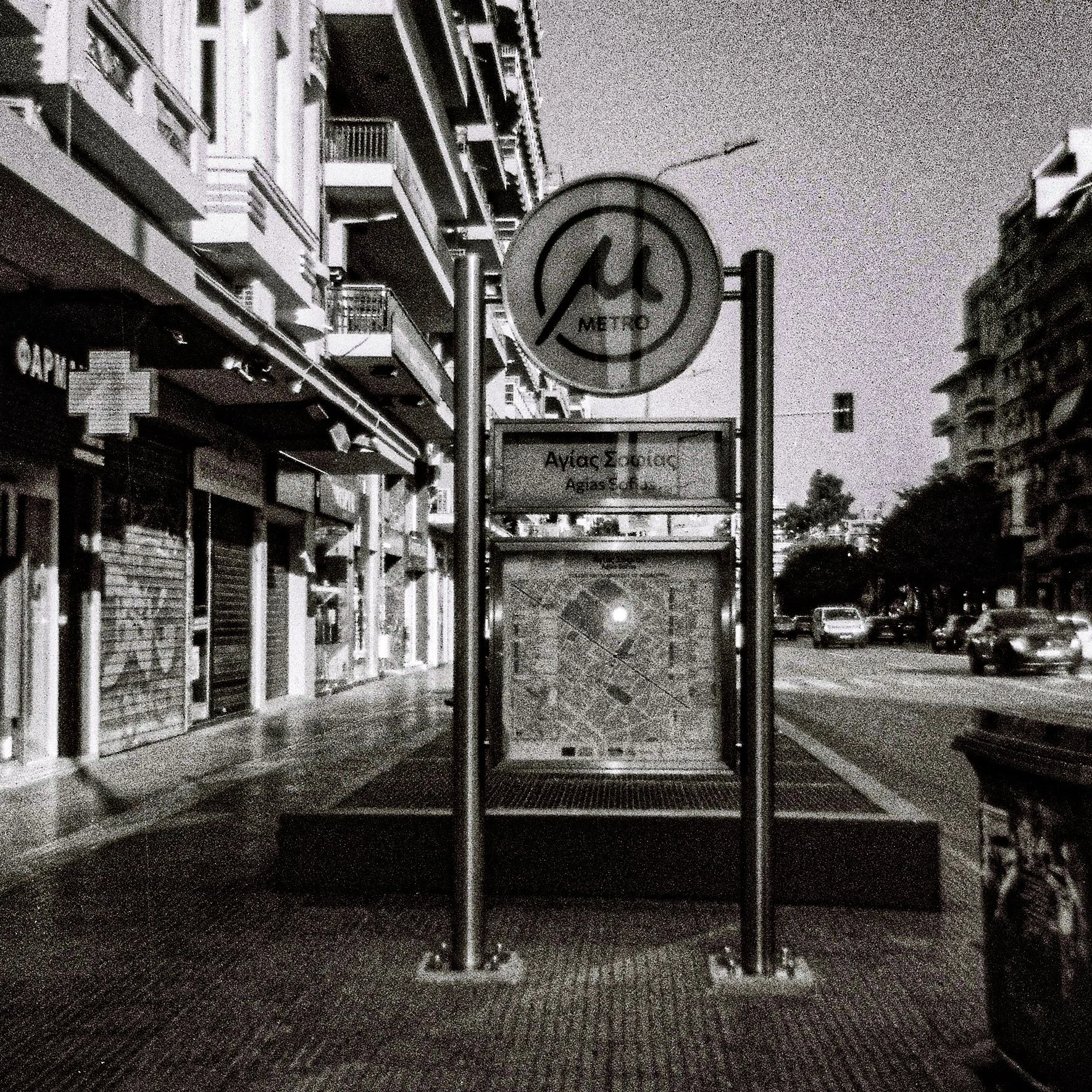 An empty city street at night with a metro sign and a bus stop in the foreground. Multi-story buildings line the street.