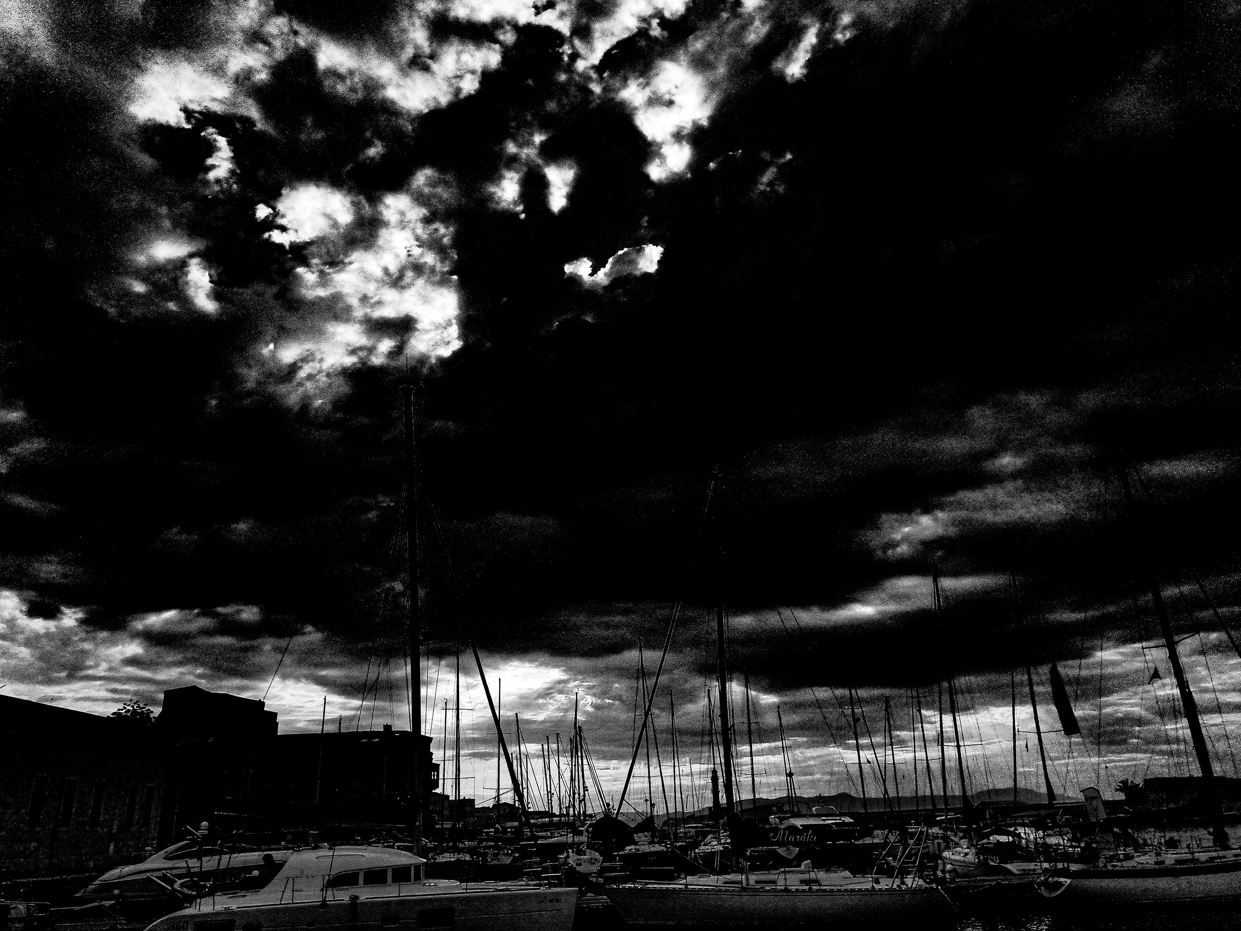 A black and white photo of a marina with boats docked, and a dramatic cloudy sky overhead.