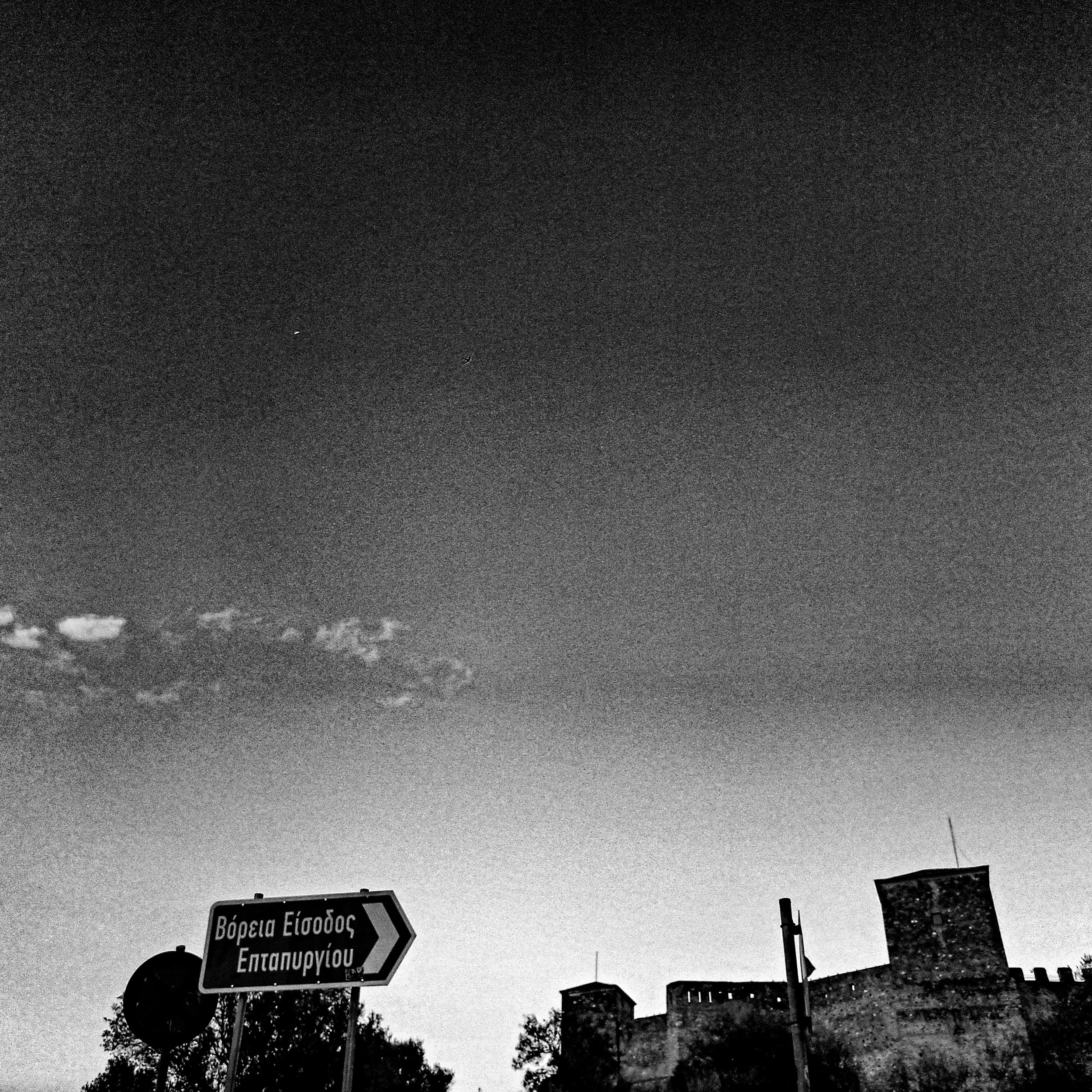 Black and white photograph of street signs with Greek text in the lower part of the image; sky filled with dark clouds; silhouettes of trees and buildings in the background.