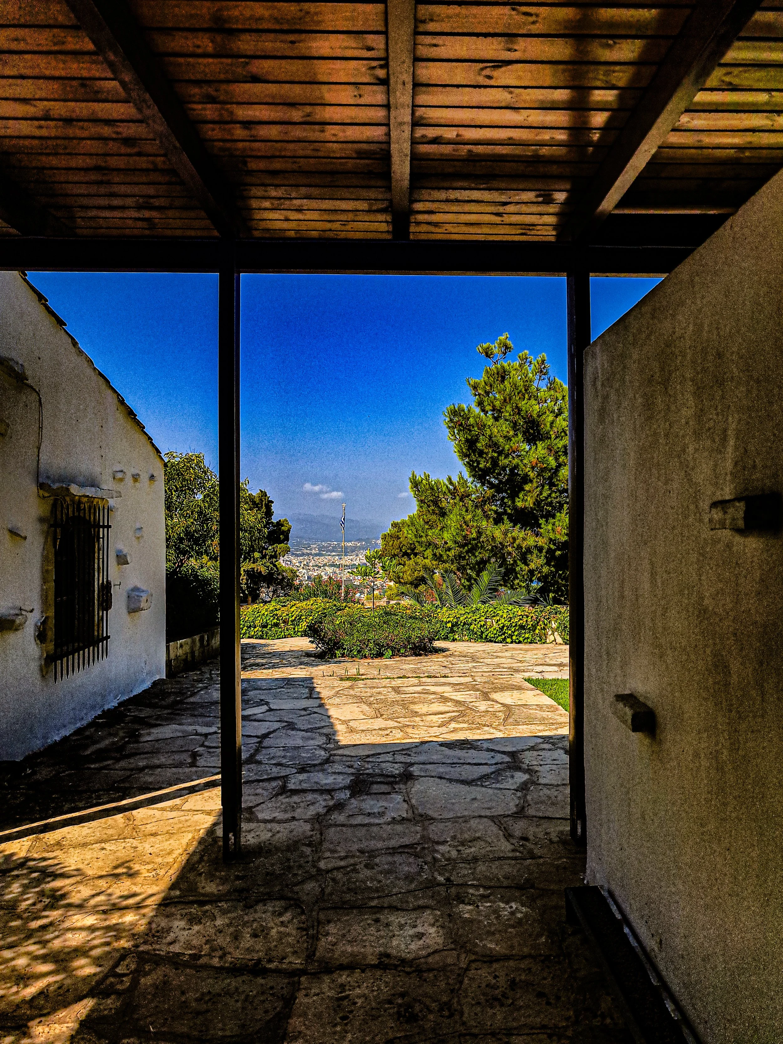 View through an open doorway towards a patio with a stone floor, trees, and a cityscape in the distance, with a tall flagpole and a bright blue sky.