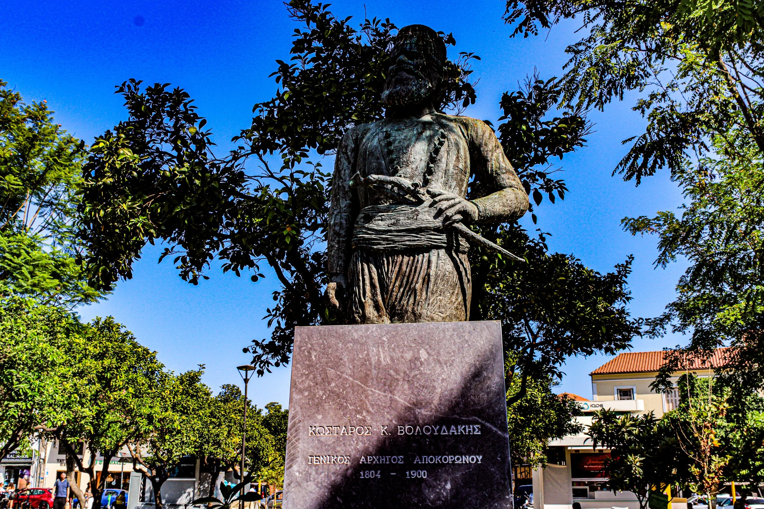 A bronze statue of a woman holding a scroll, standing on a stone pedestal in a park surrounded by trees and buildings, under a clear blue sky.