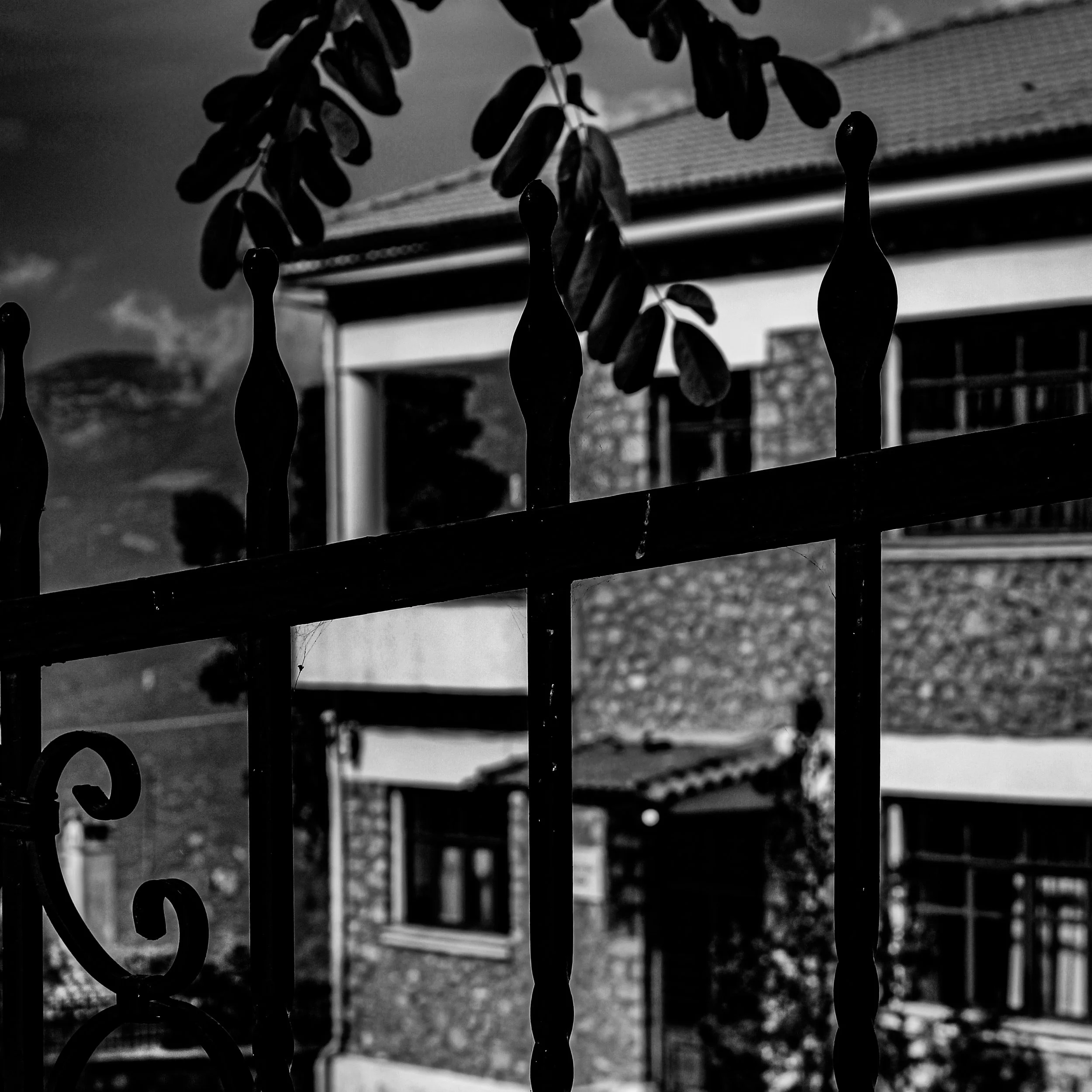 Black and white photo of a house viewed through a decorative metal fence with ornate design and vertical bars. The house has a triangular roof and a window, and the sky is cloudy.
