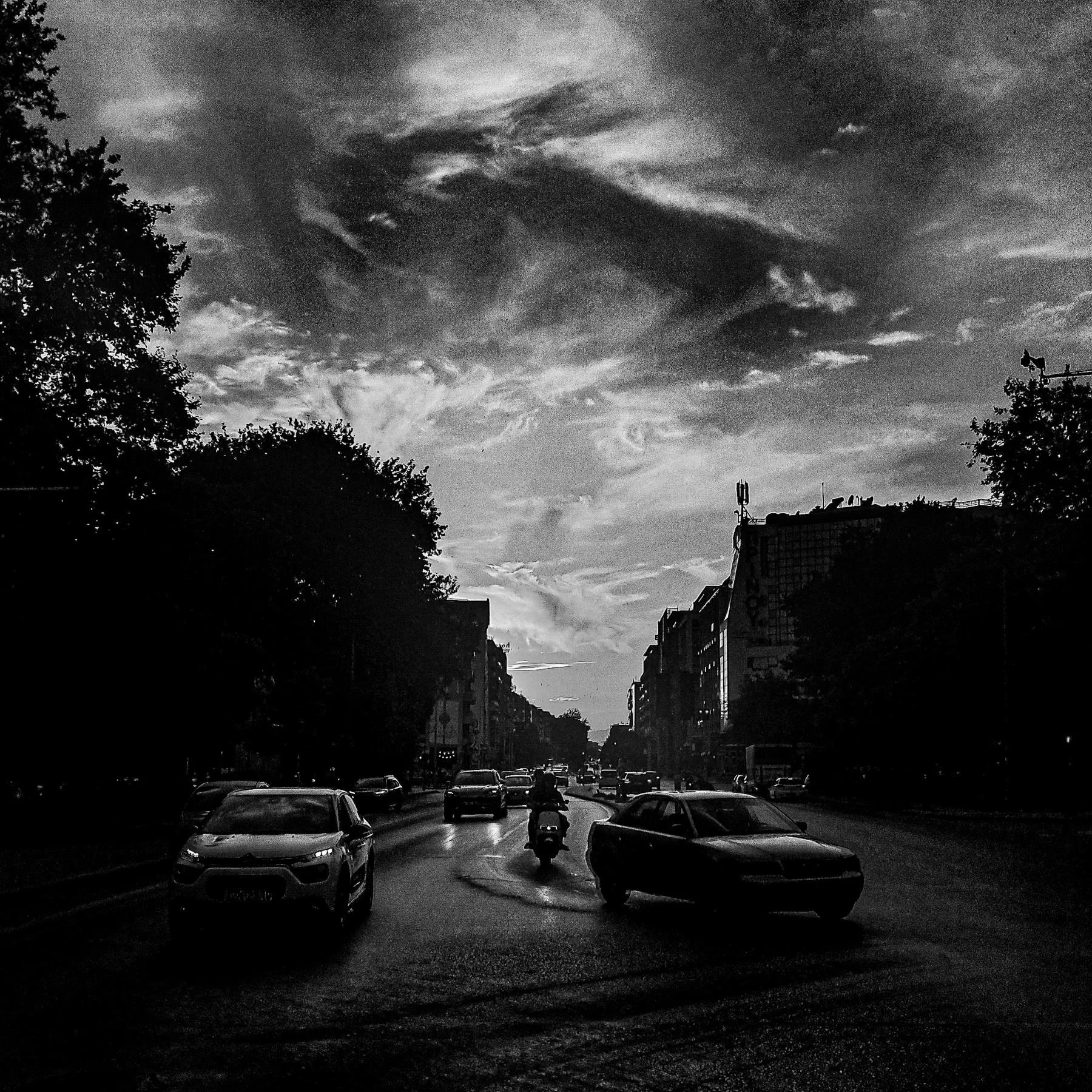 Black and white photo of a city street with cars and a motorcycle riding in the middle. Tall buildings and trees line the street. The sky is overcast with clouds, creating a moody atmosphere.