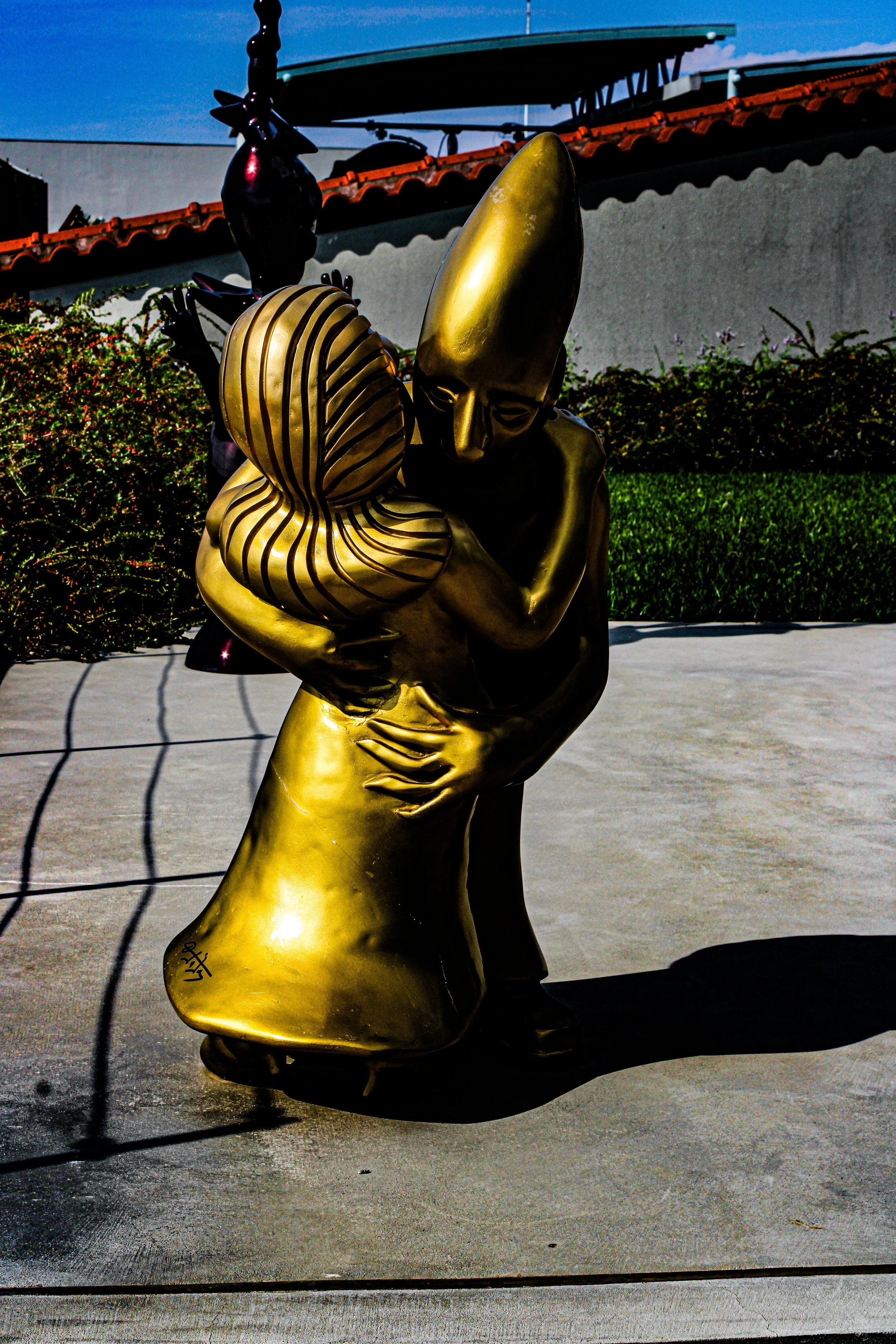 A gold sculpture of a person holding a child, with a metallic black rabbit figure positioned behind them, outdoors on a sunny day with a building, greenery, and a blue sky in the background.