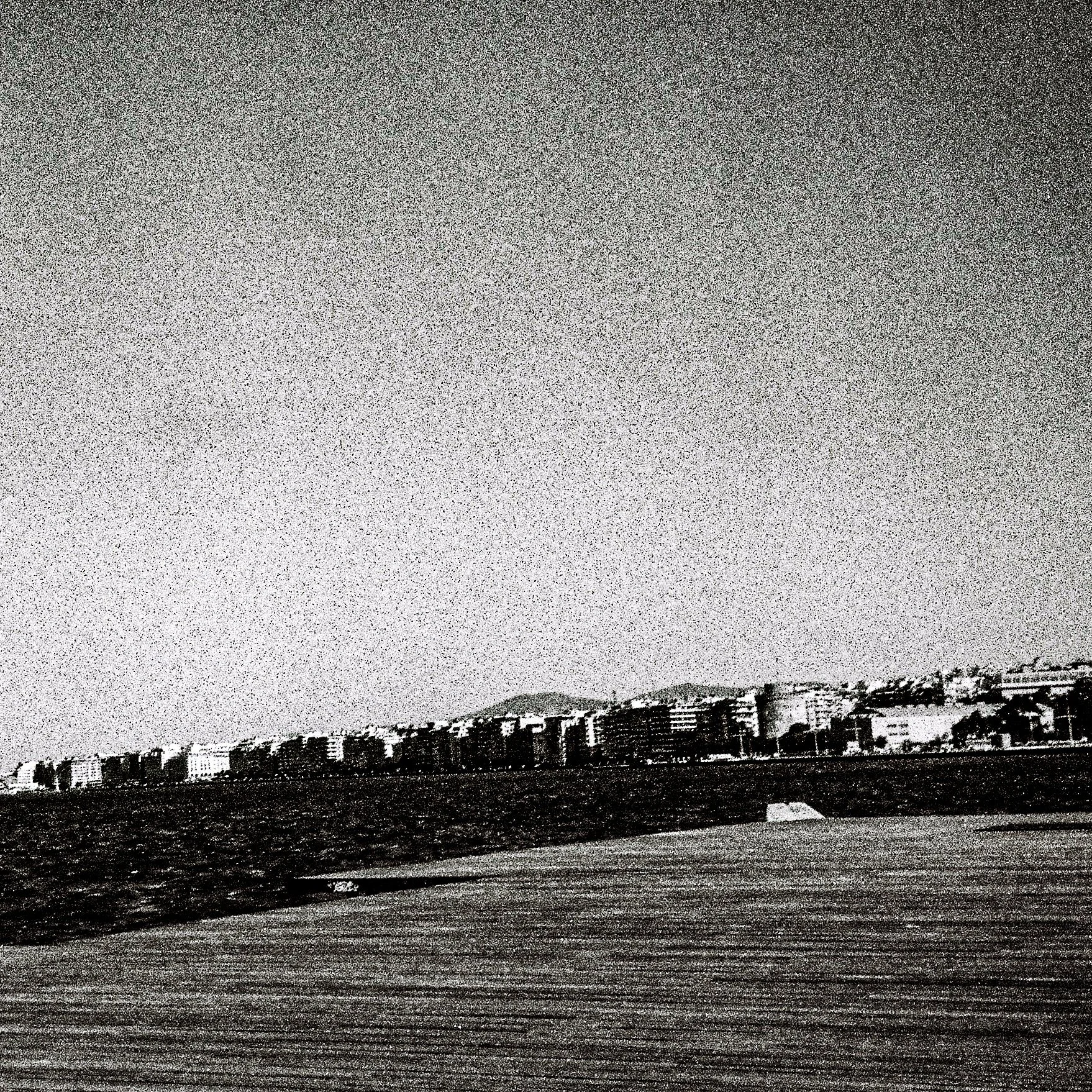 Black and white photo of a cityscape with buildings along the coastline and a cloudy sky.