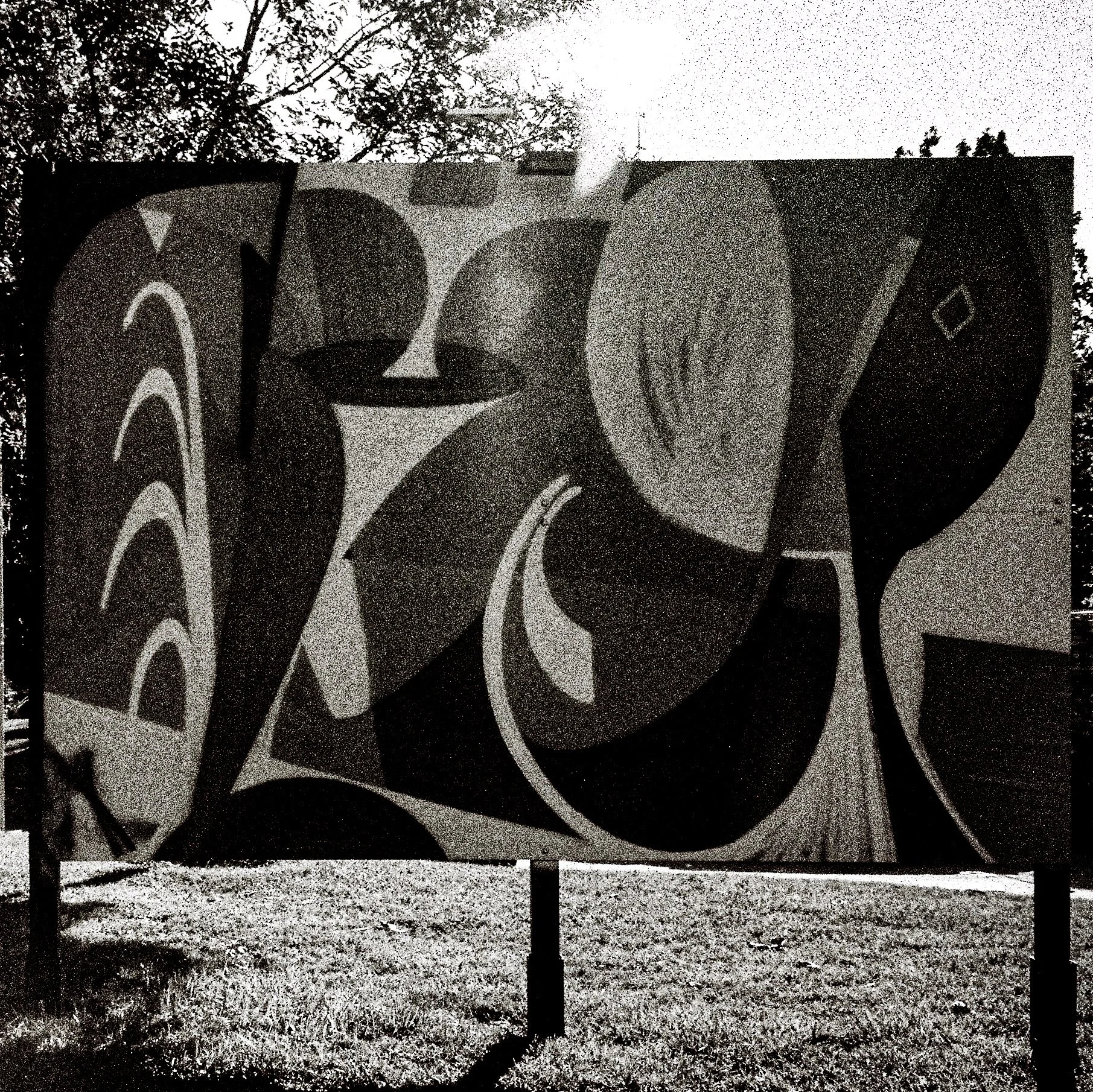 Black and white graffiti mural with stylized, abstract letters on a wall, outdoors near trees and a small fenced area.