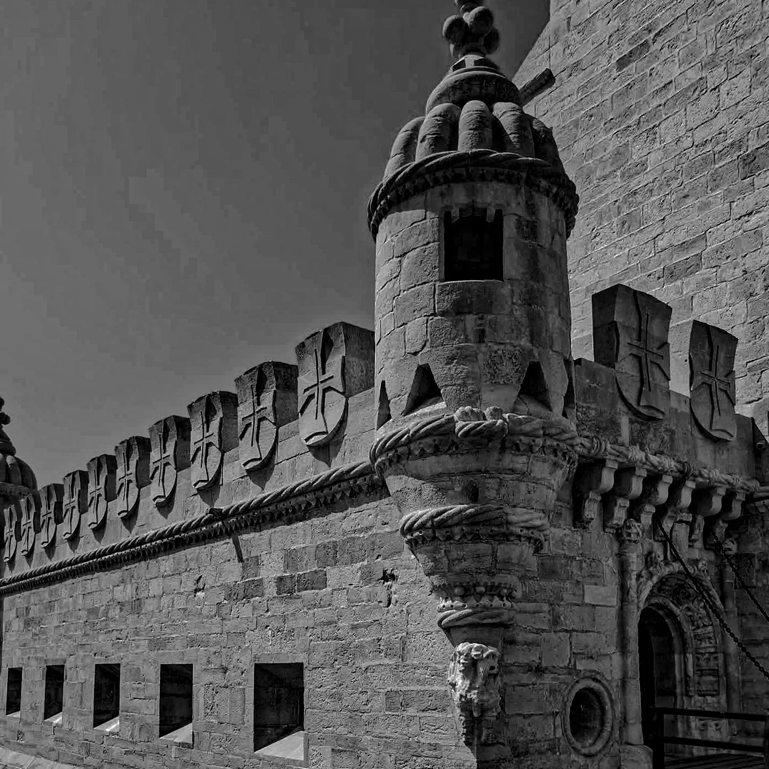 Black and white photograph of a historic stone building featuring a decorative turret with a small arched window, ornamental battlements with shield-shaped carvings, and a carved lion head sculpture near an arched doorway.