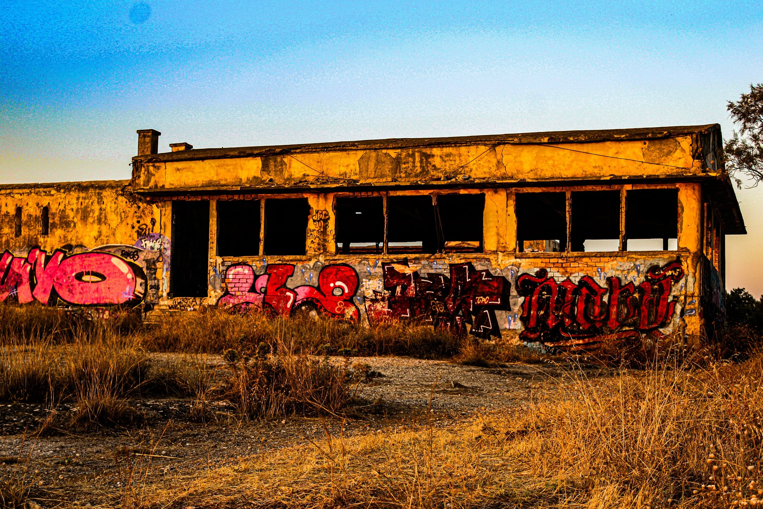 An abandoned, multi-story building with yellow, peeling paint, graffiti on its walls, and open windows, surrounded by dry grass under a clear blue sky.
