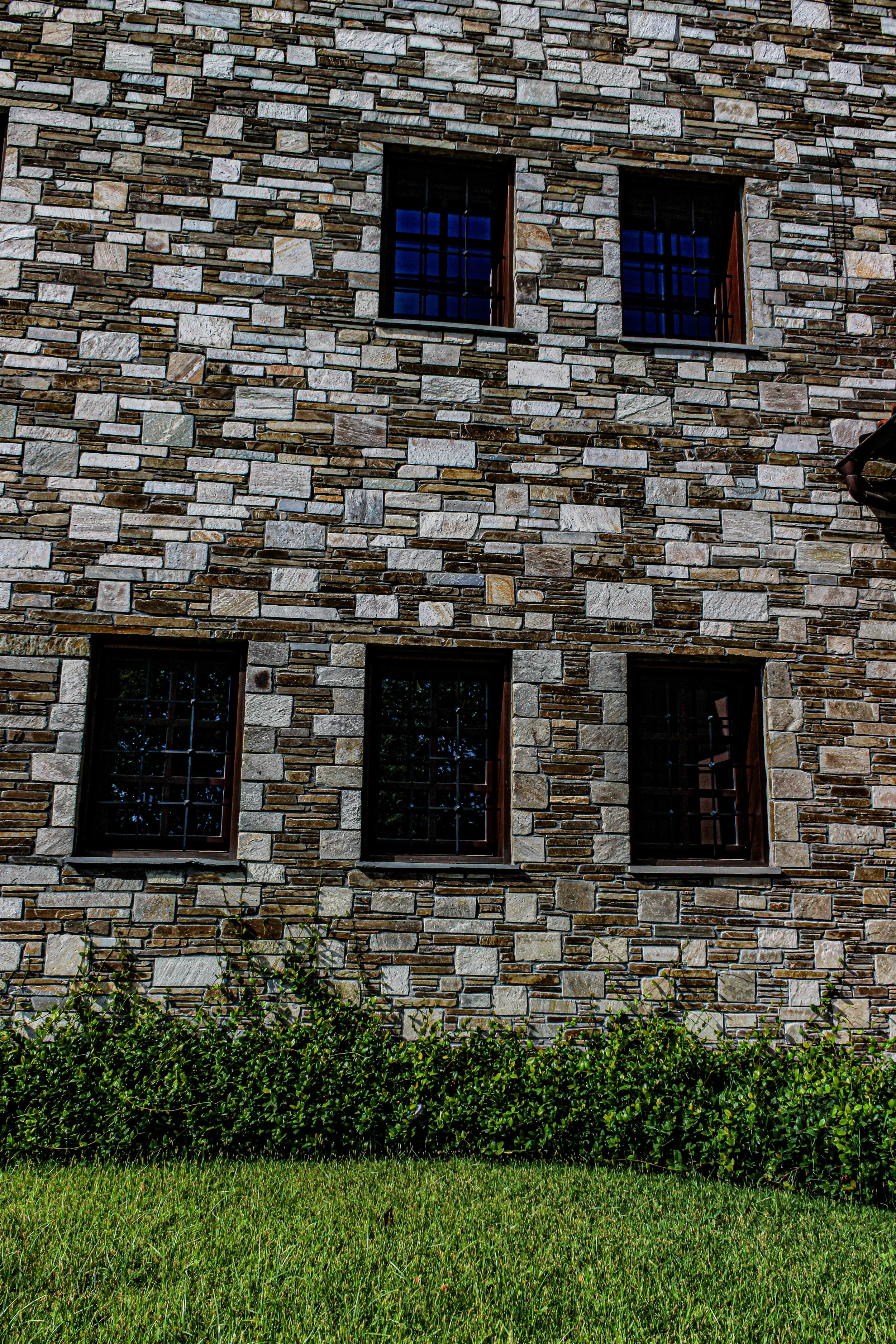 A stone brick wall with three small windows, complete with black metal bars, and greenery including grass and bushes at the base.