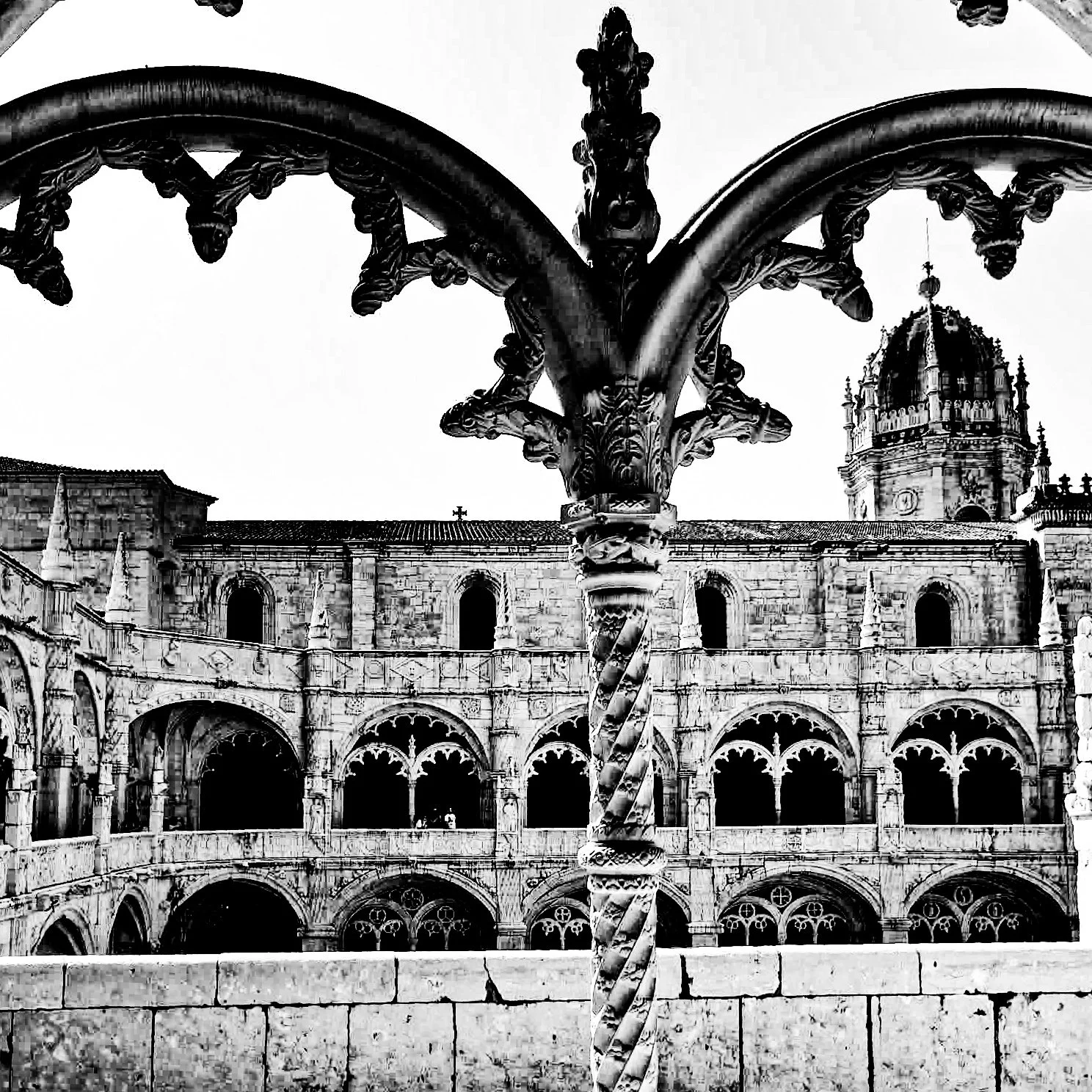 Black and white photo of a twisted metal column in an outdoor courtyard with arches and a building with domed tower in the background.
