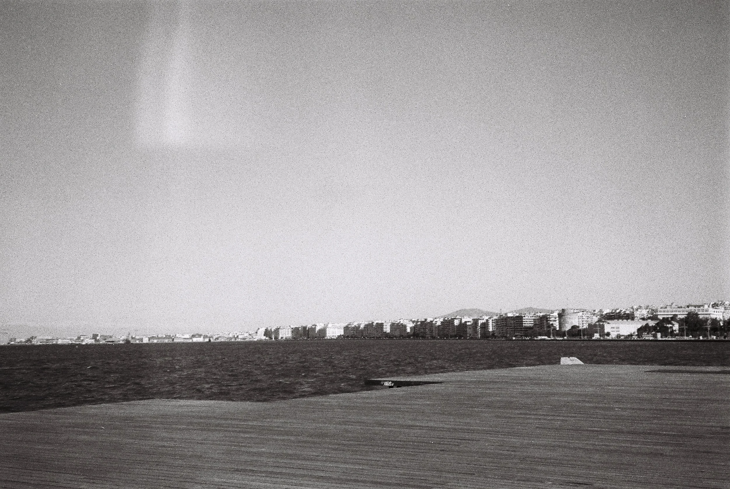 Black and white photo of a coastal cityscape with multiple buildings along the shoreline, with a large body of water in the foreground and mountains in the background.