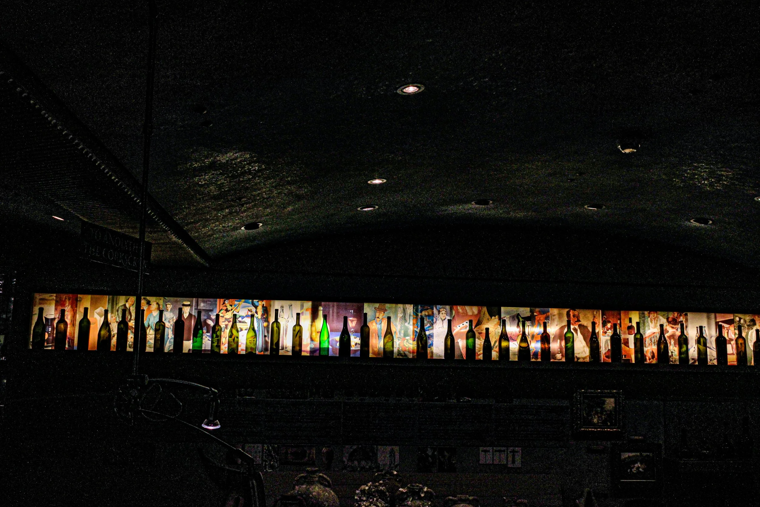 A dark interior with a row of illuminated bottles of various colors in front of a colorful mural or advertisement.