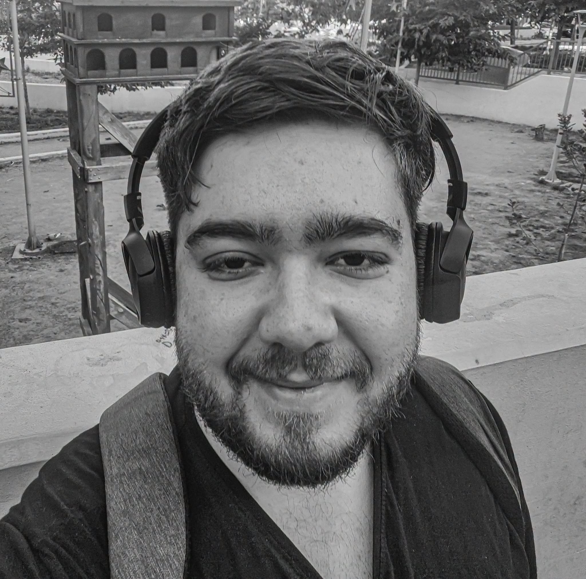 A person with dark hair and a beard wearing large headphones. The photo is in black and white, taken outdoors in a park or garden area with trees and a wooden structure in the background.