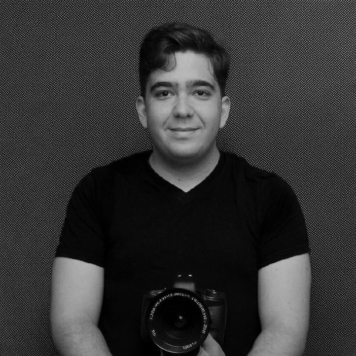 A young man takes a selfie with a digital camera, wearing a black t-shirt, standing against a textured background.