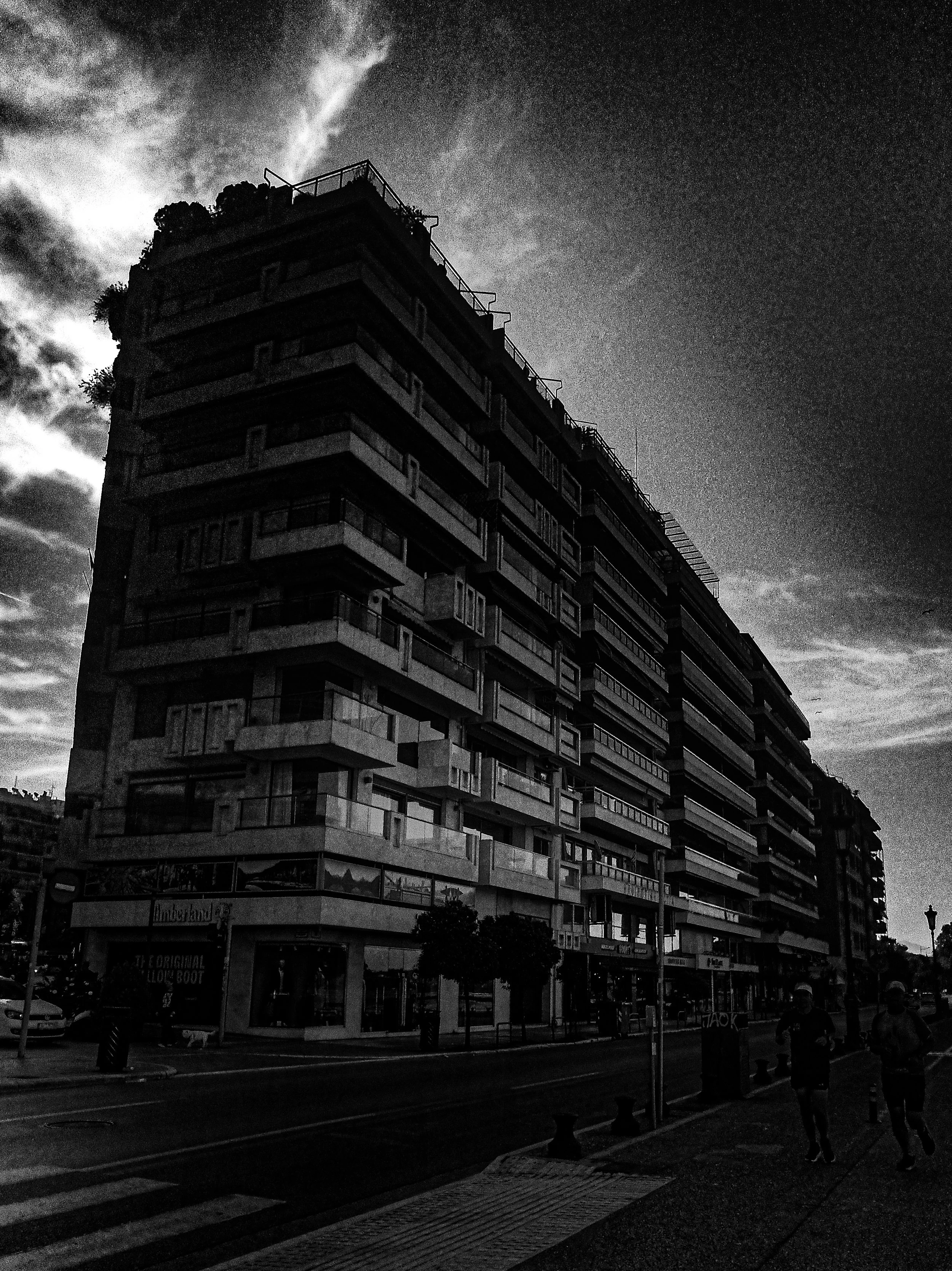 A black and white photo of a tall modern building with multiple balconies and large windows, taken from street level with a few pedestrians and vehicles, and cloudy sky above.