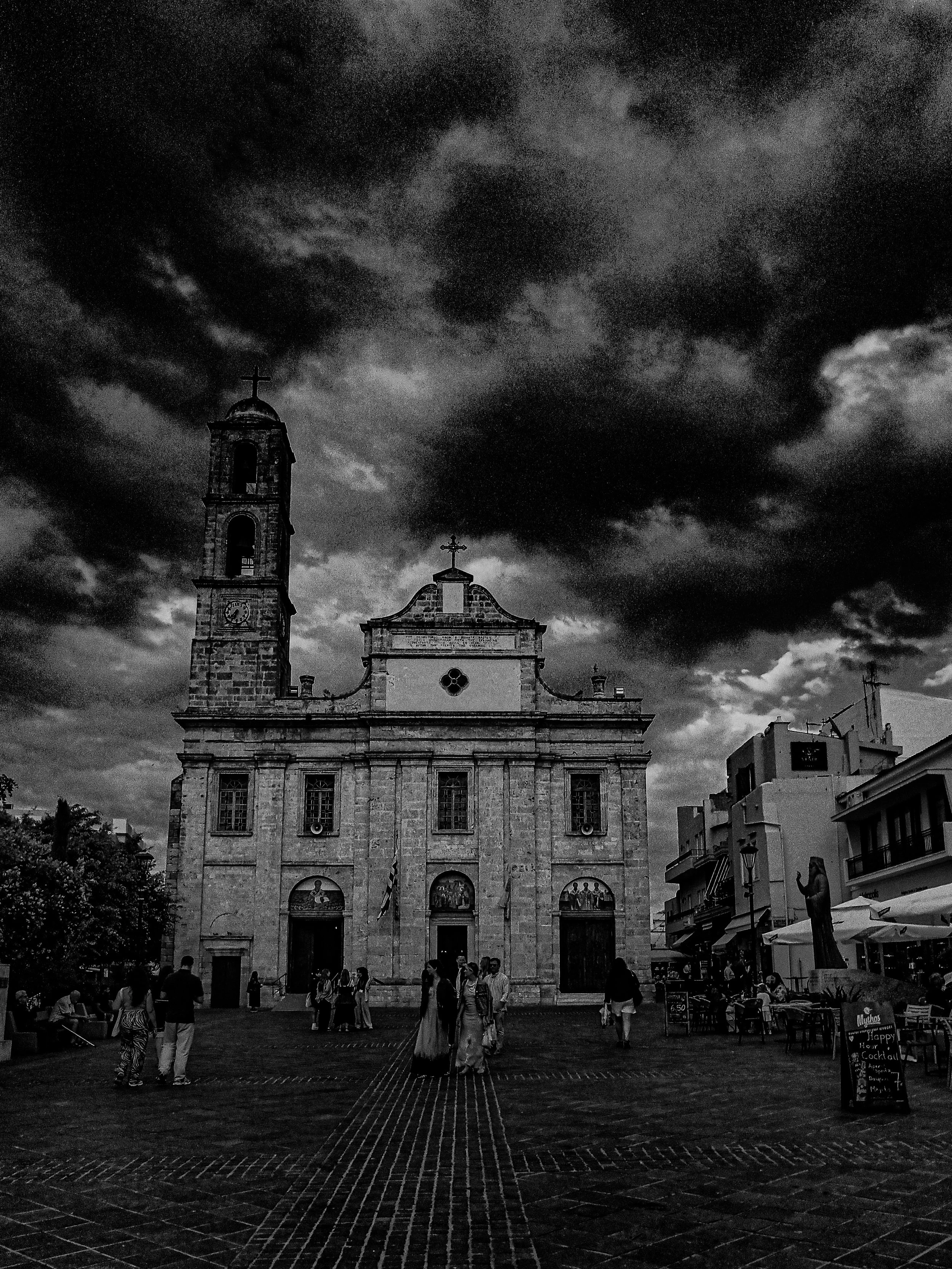 A black and white photo of a historic church with a tall bell tower topped with a cross, situated in a bustling town square filled with people. The sky above is cloudy and dark, creating a moody atmosphere.