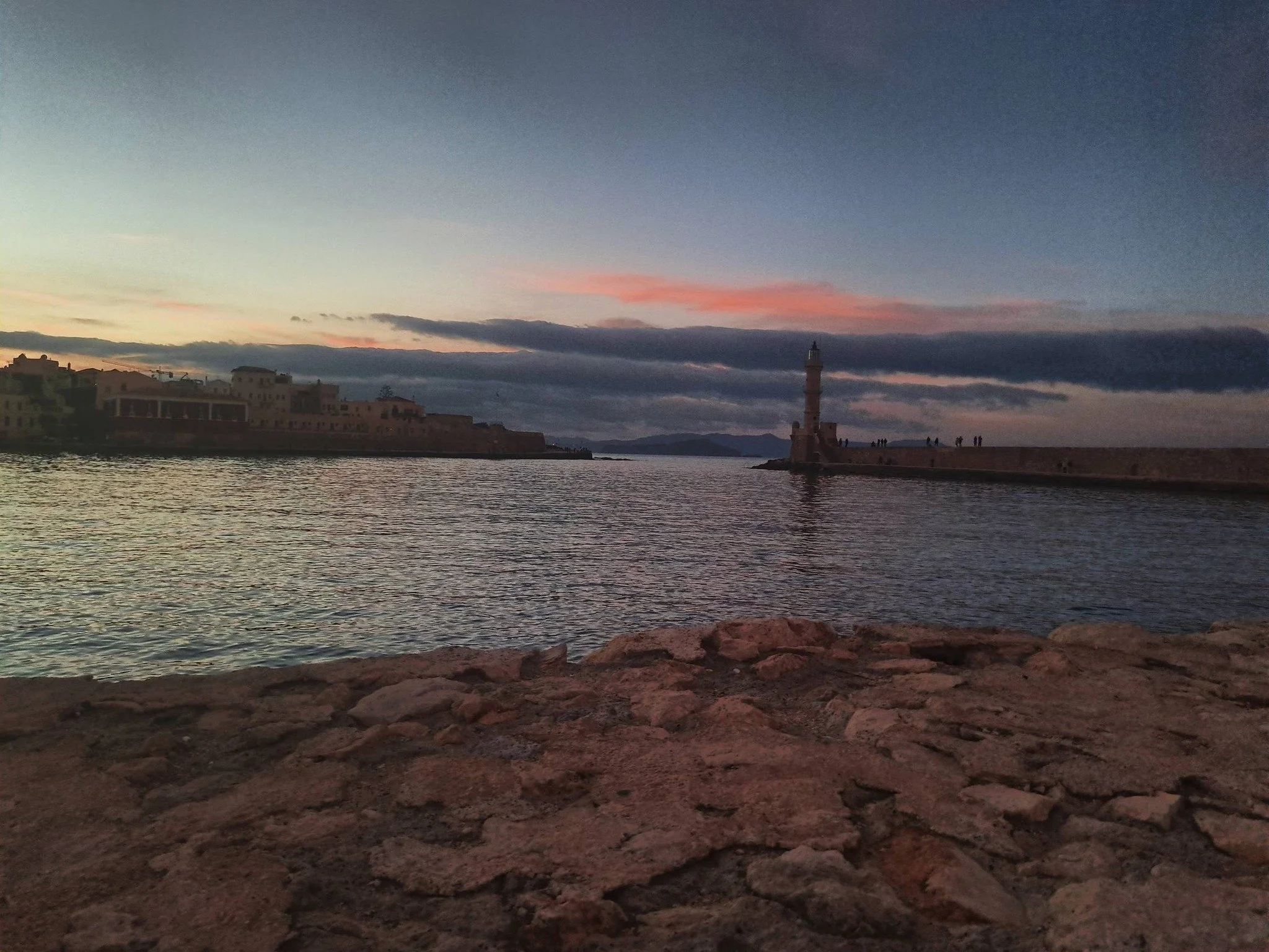 A sunset over a harbor with buildings on the left and a lighthouse on the right, calm water reflecting the sky, and people walking along the harbor wall.