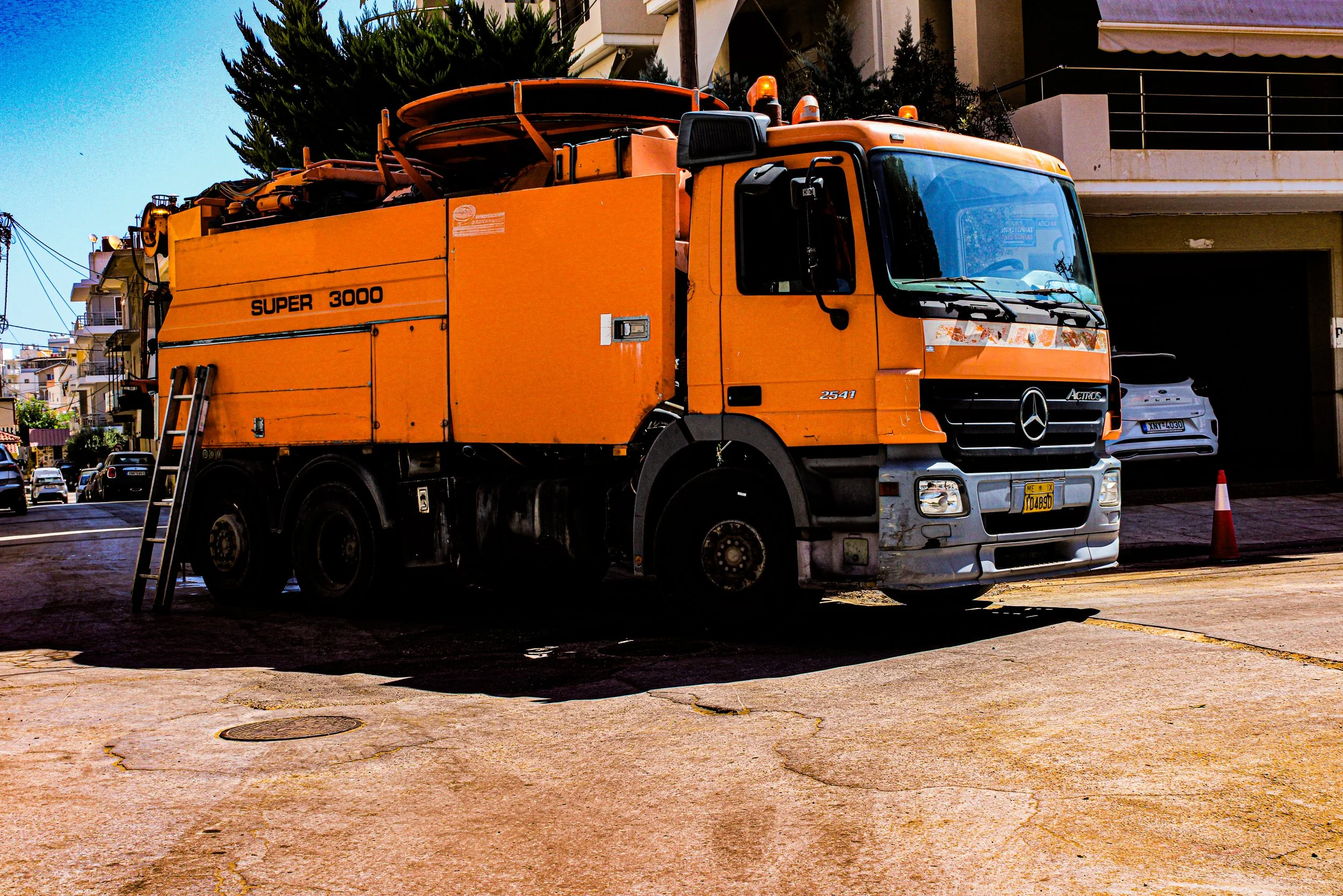 An orange utility truck parked on a city street, with a ladder leaning against it and a white car parked nearby. Residential buildings with balconies and trees are in the background.