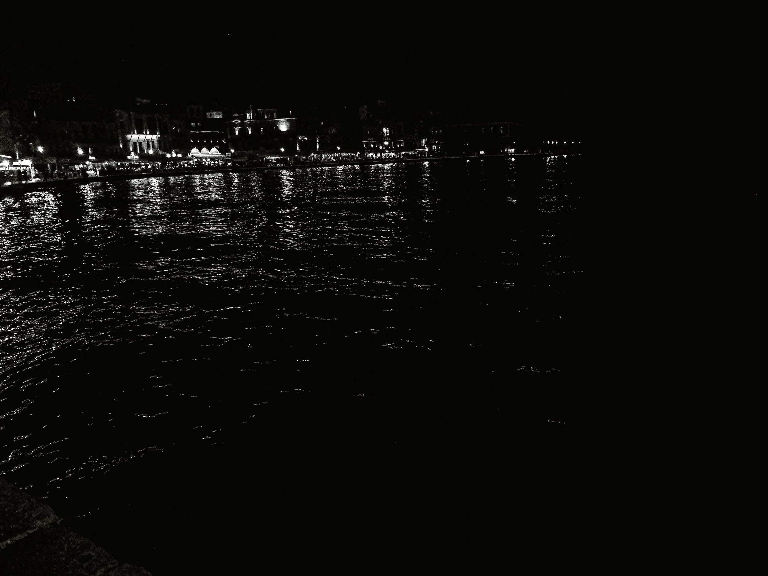 Nighttime scene of a waterfront or harbor with buildings and lights reflected on the water.