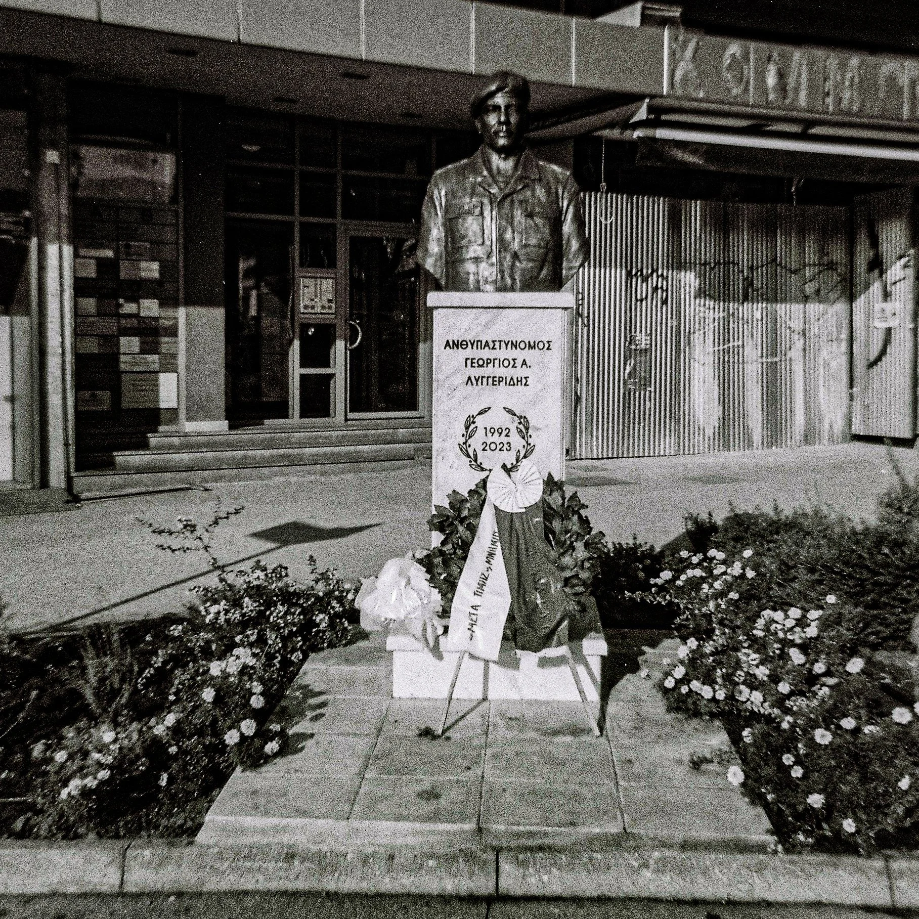 Memorial monument with a bronze bust of a man wearing a jacket, set on a granite pedestal. The pedestal has Greek inscriptions. Flowers and wreaths are placed at the base. The background shows a glass entrance door and a metallic wall.