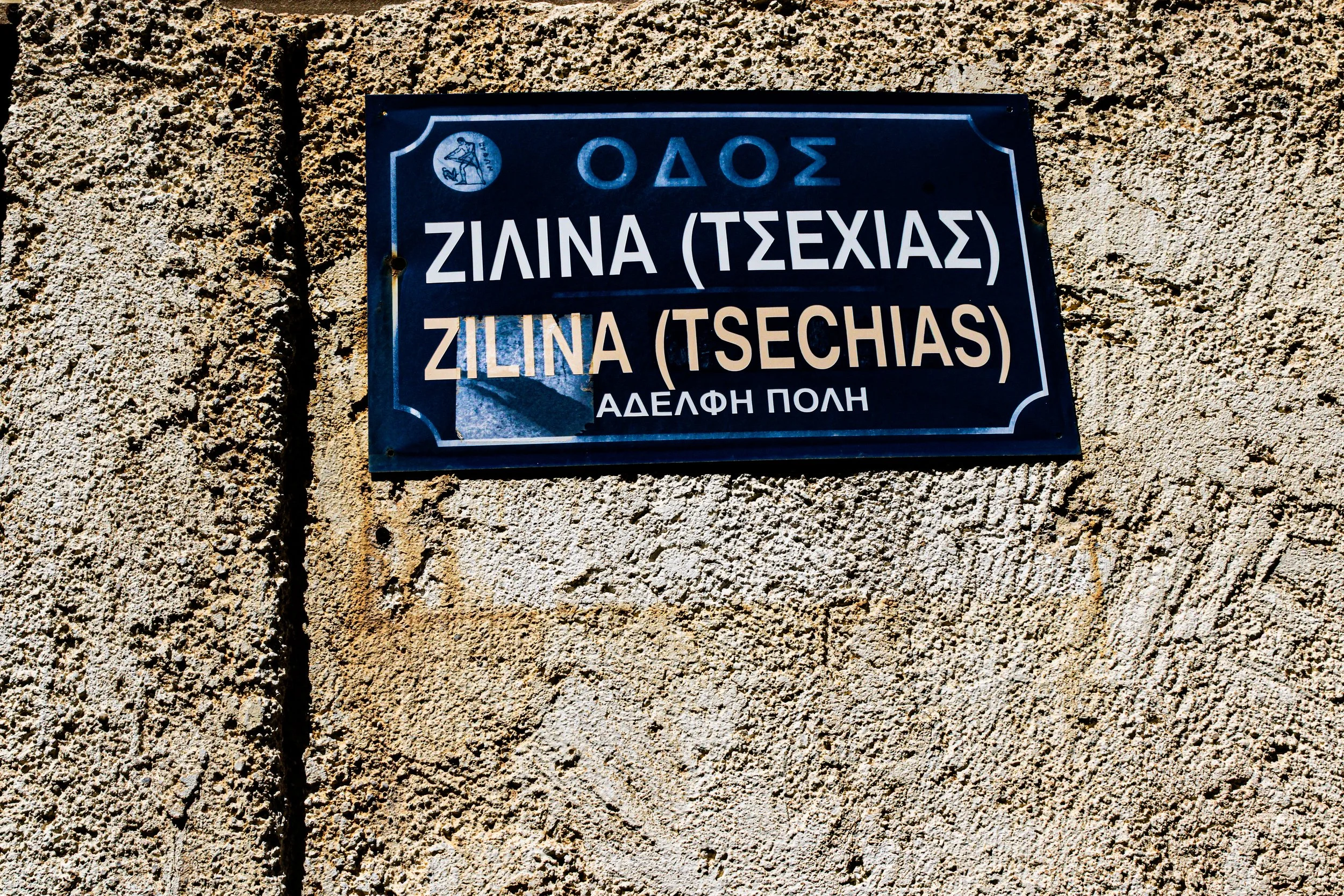 Street sign in Greek on a textured stone wall. The sign has an emblem in the upper left corner and reads, 'Odos Zilina (Tsehias)'. Small holes are visible in the sign.