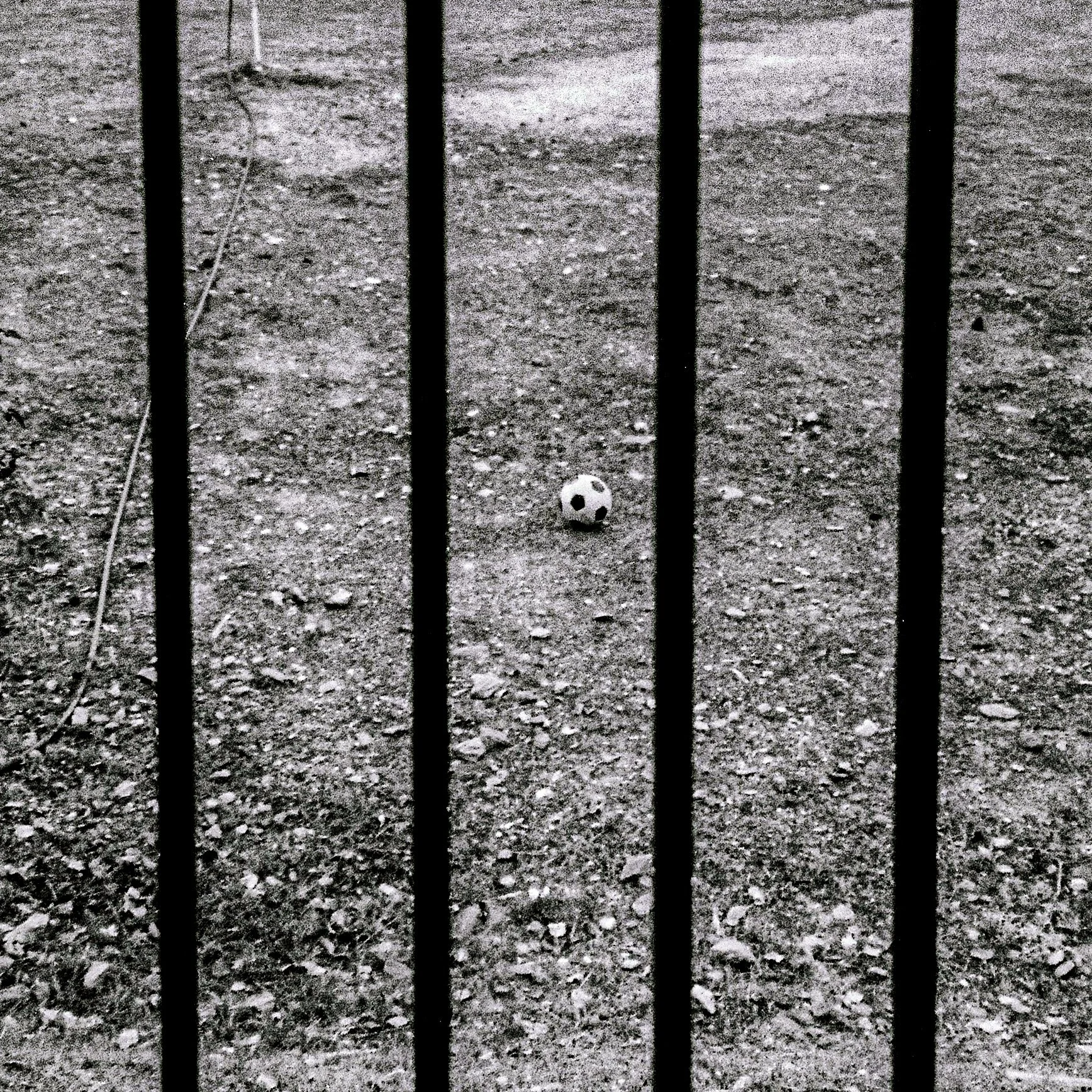A small black and white soccer ball on the ground behind metal bars, as seen through a fence, in a gravel or dirt area.