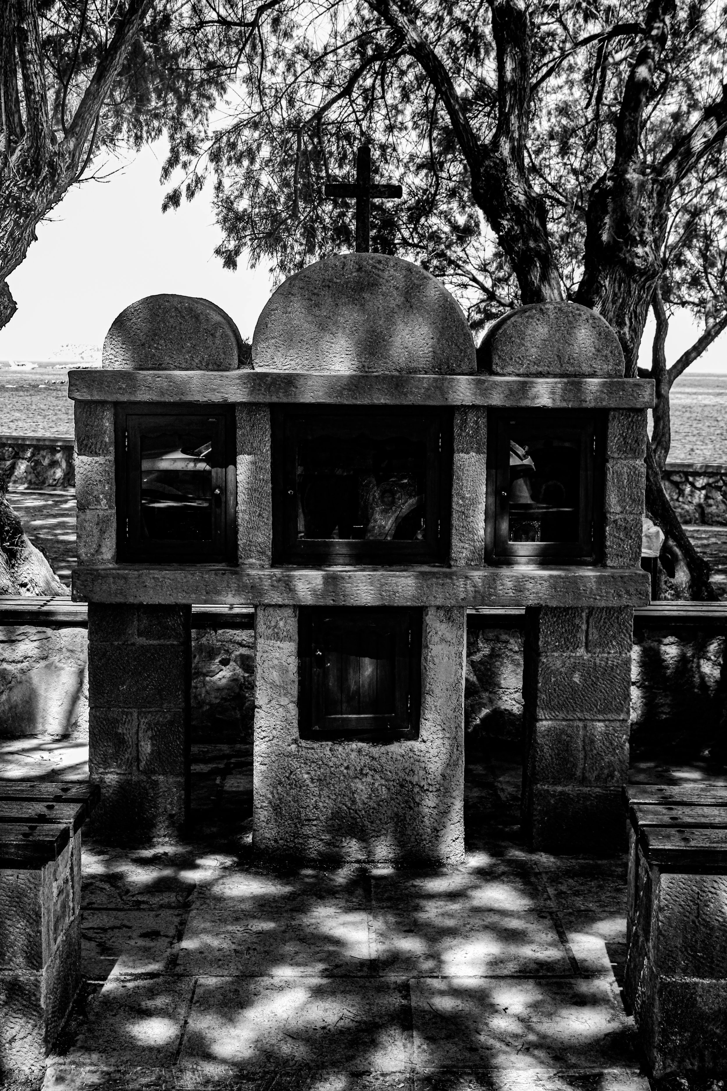 Black and white photo of a small outdoor shrine with a cross on top, set between two trees, with shaded ground and pathways leading to it.