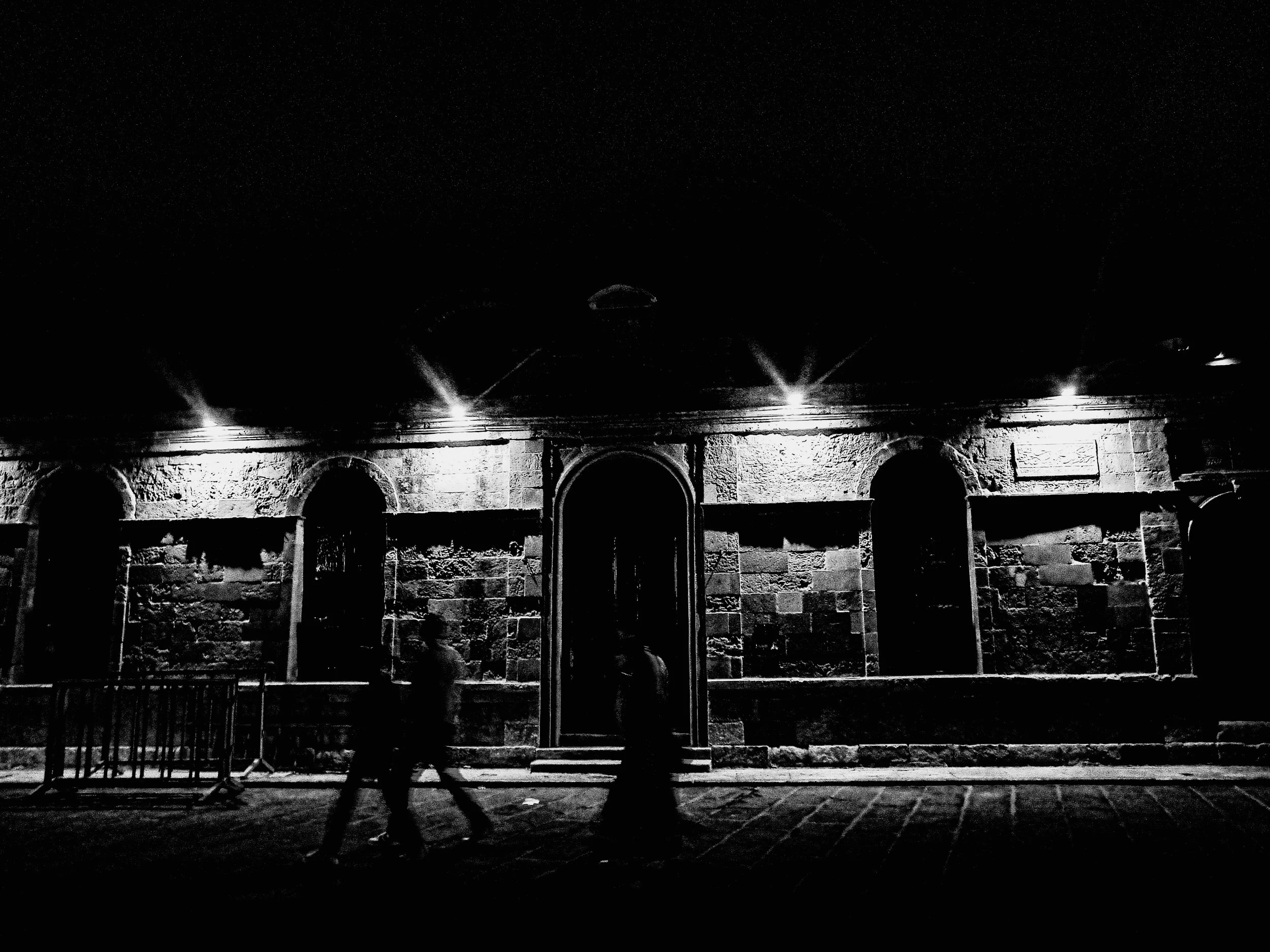 Night view of a historic stone building with arches, illuminated by streetlights, and a person walking in front.