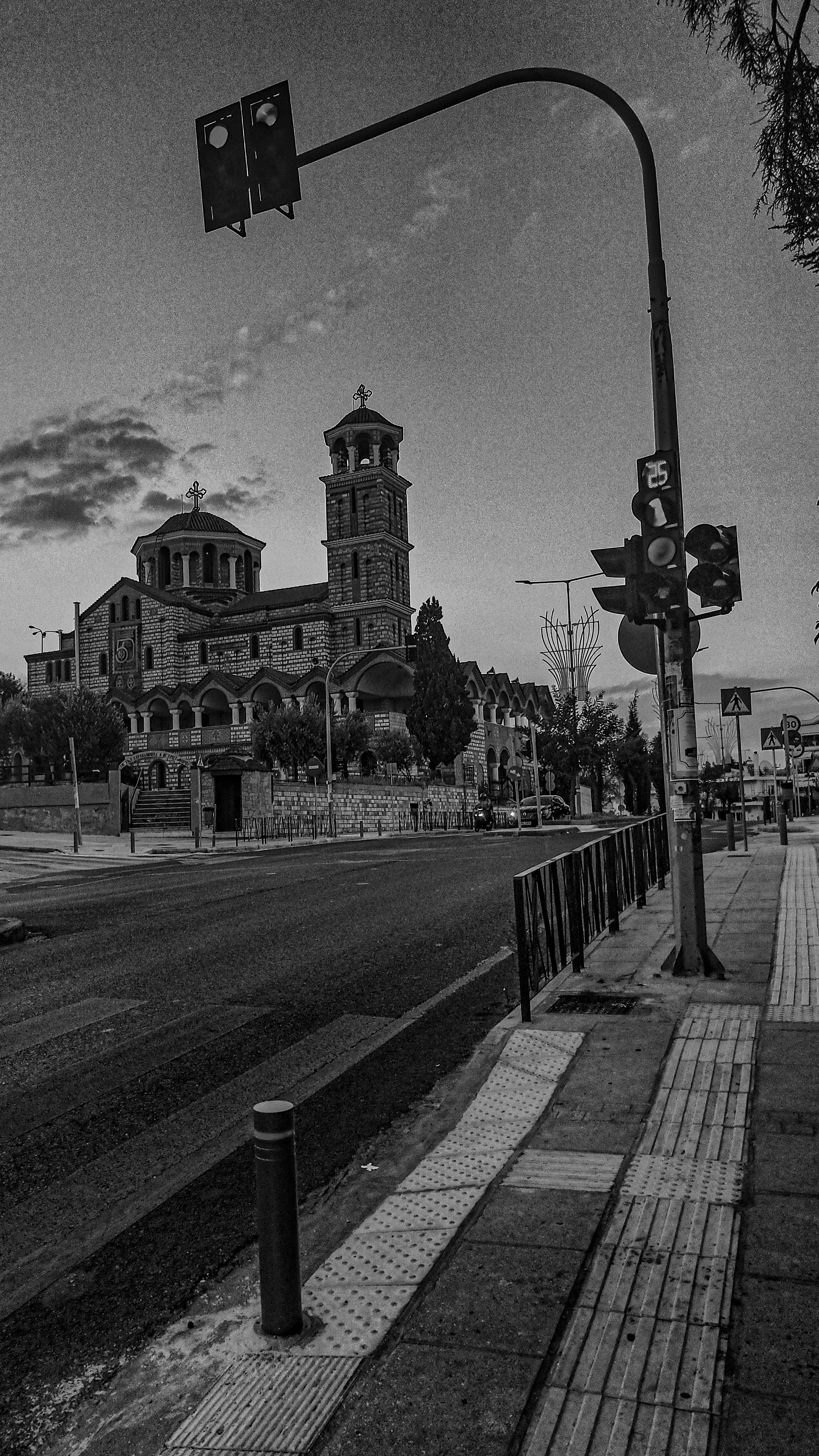 A black and white photo of a church with two bell towers, streetlights, traffic lights, a cross on top of both towers, and a sidewalk with a textured tactile paving segment.