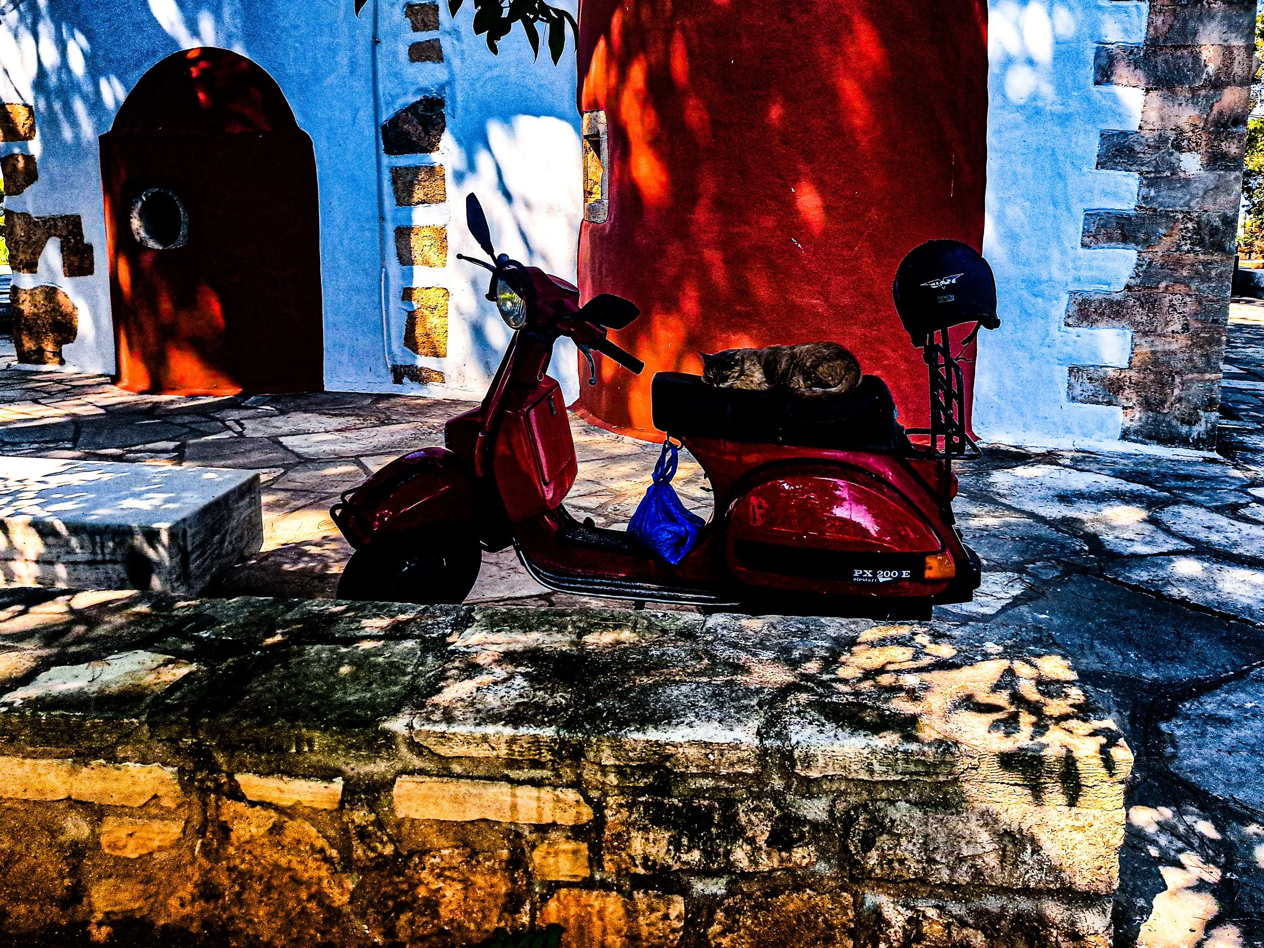 A red scooter parked in front of a colorful painted wall with a cat resting on the scooter seat.