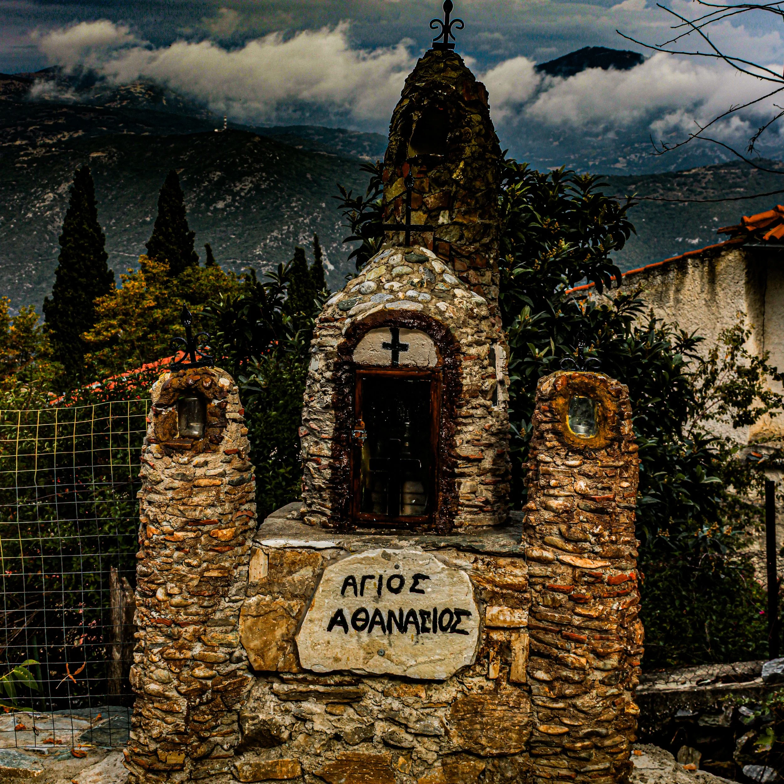 A stone structure with a cross on top, set against a mountainous landscape with cloudy skies. There is a sign with Greek writing at the bottom of the structure.