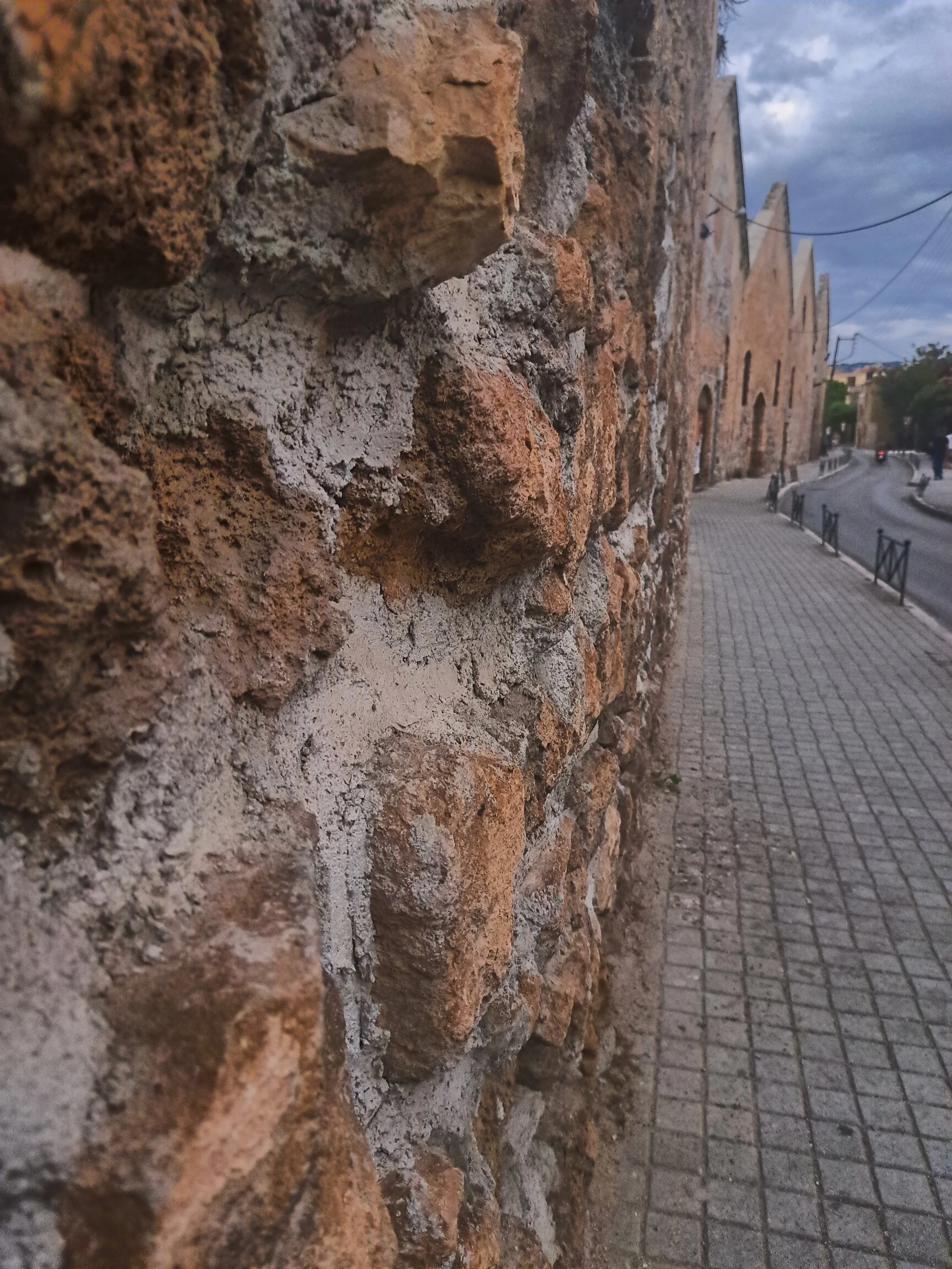 Close-up view of an old brick wall with a sidewalk and road in the background, during cloudy weather in an urban setting.