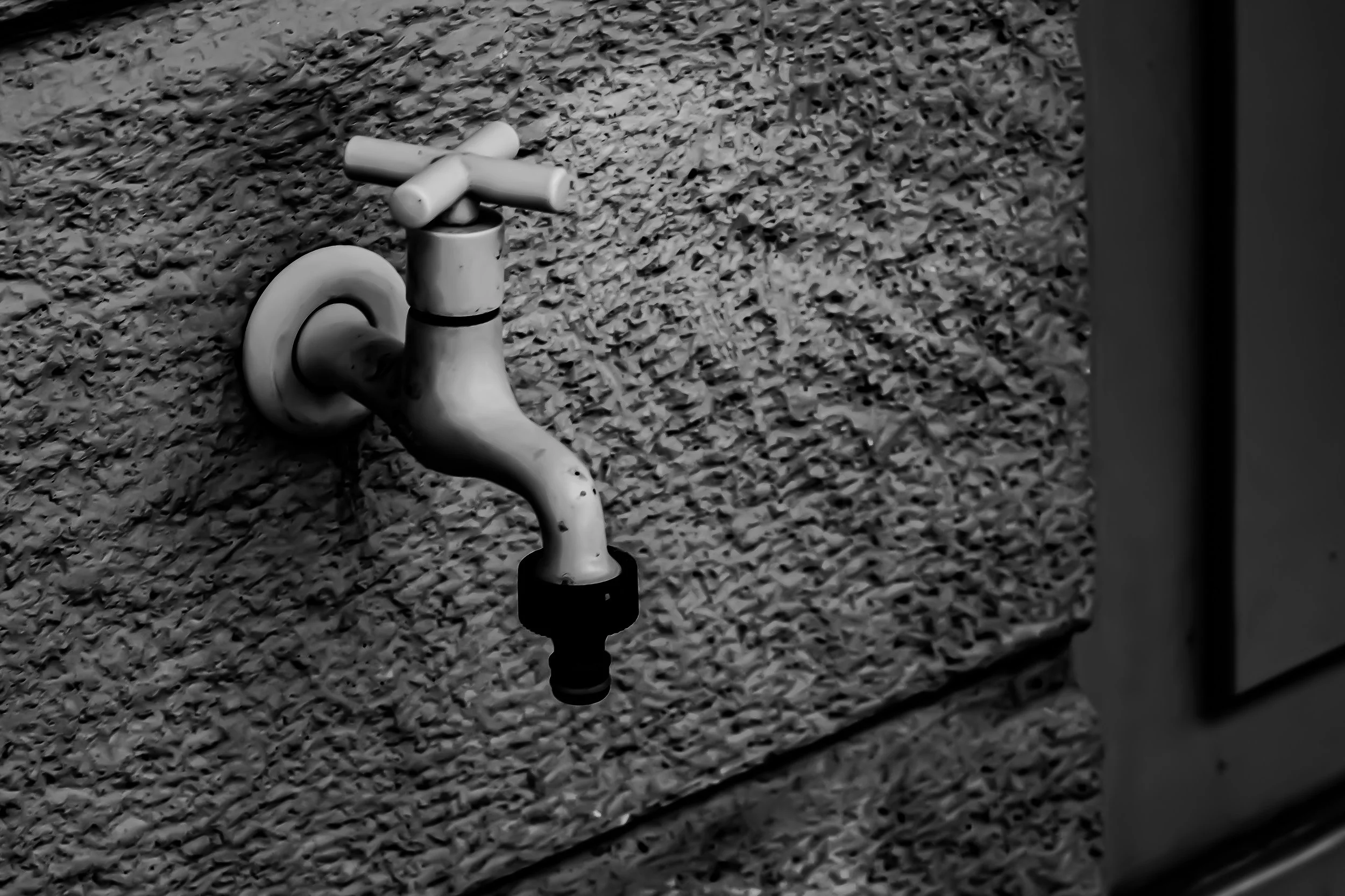 Black and white photograph of a faucet with a black hose attached, positioned outdoors on a gravel surface.