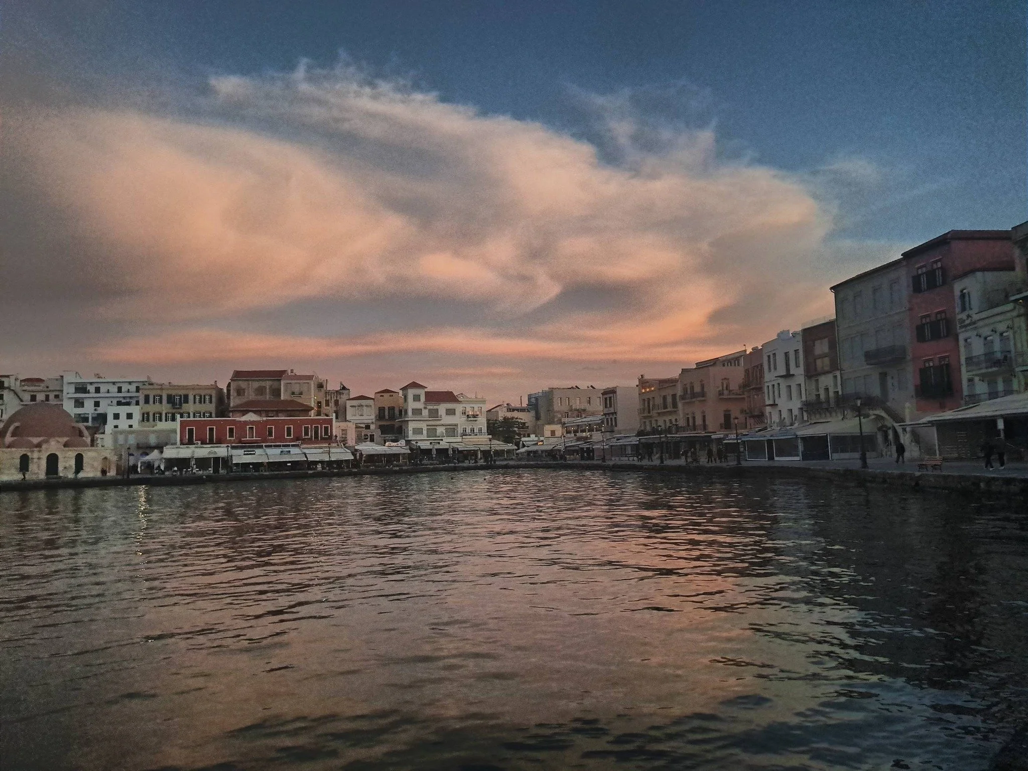 A cityscape at dusk with pastel-colored clouds in the sky, buildings lining a waterfront, and calm water reflecting the scene.