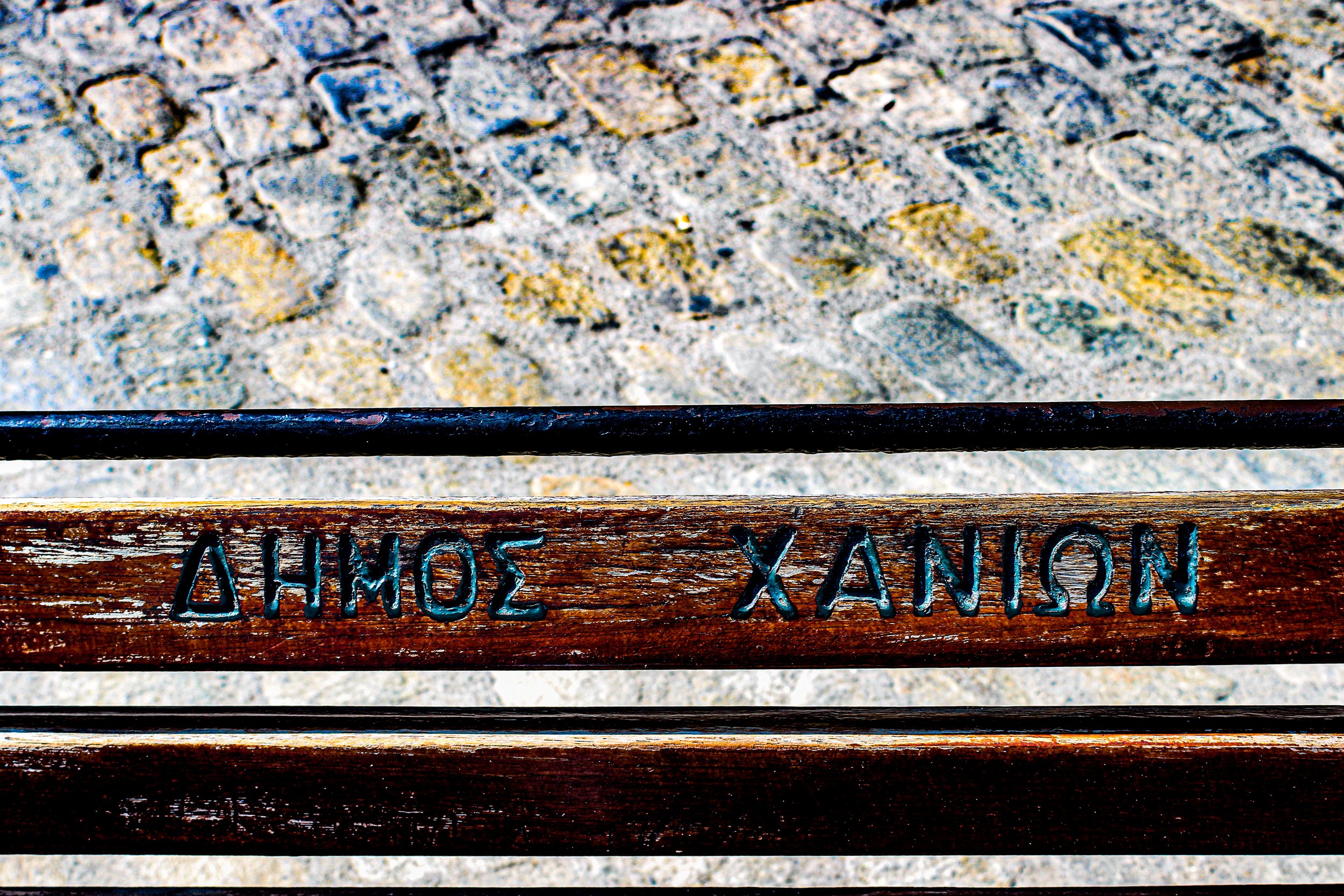Close-up of a wooden bench with metal letters that spell out "XANHÔZ" and part of metal railings above it. The background contains cobblestone pavement.