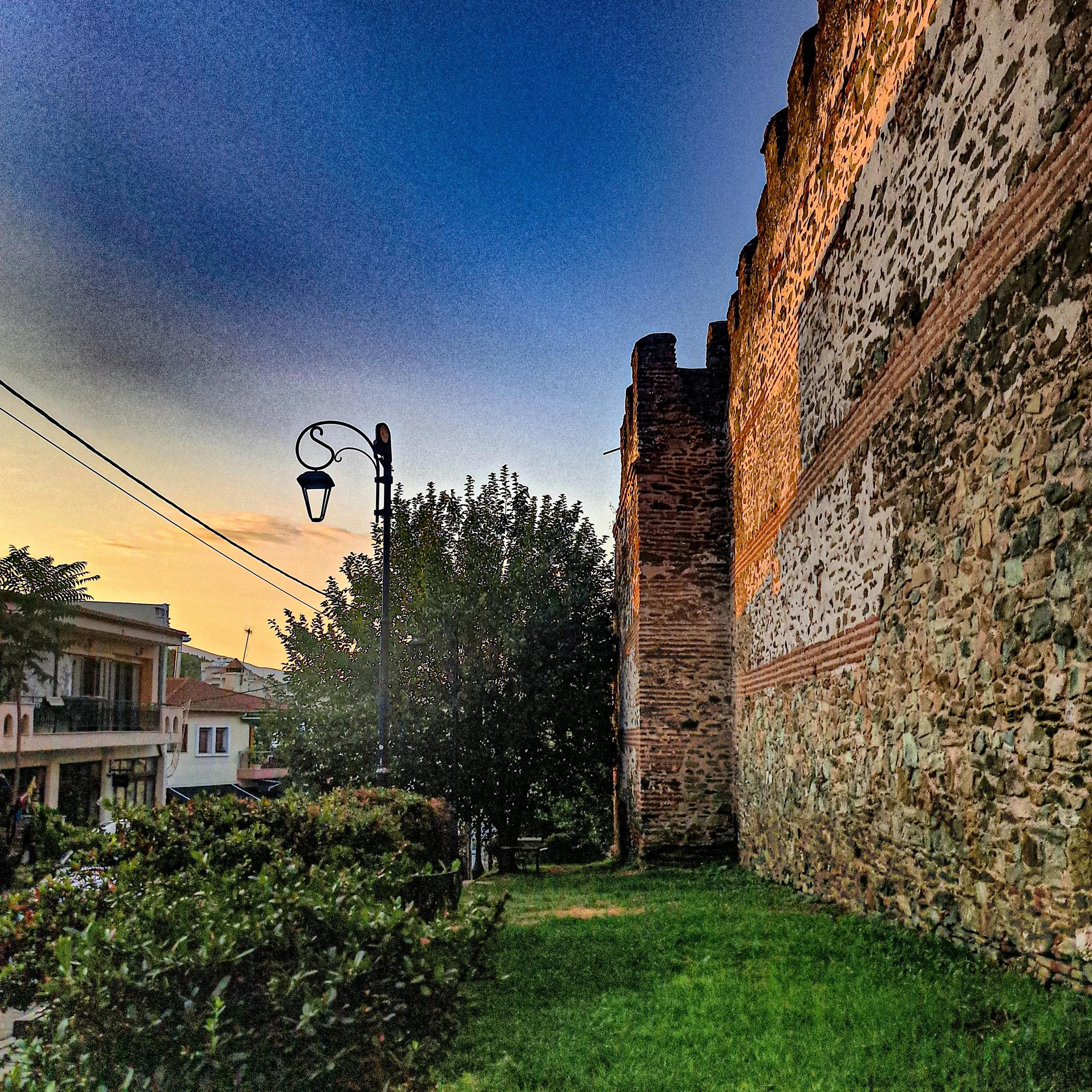 Historical brick and stone wall with chimney, green lawn, trees, and residential buildings in background during sunset.