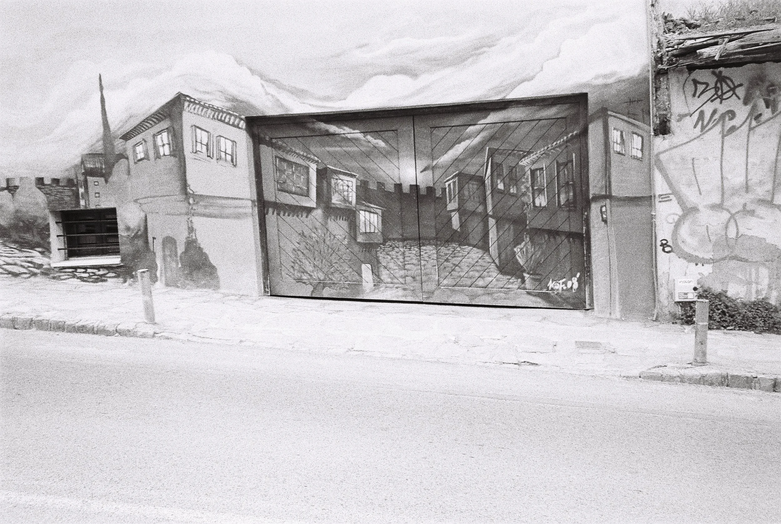 Black and white photo of a mural on a wall depicting buildings and a courtyard, with graffiti and graffiti tags on the wall, and a parked vehicle partially visible on the left.