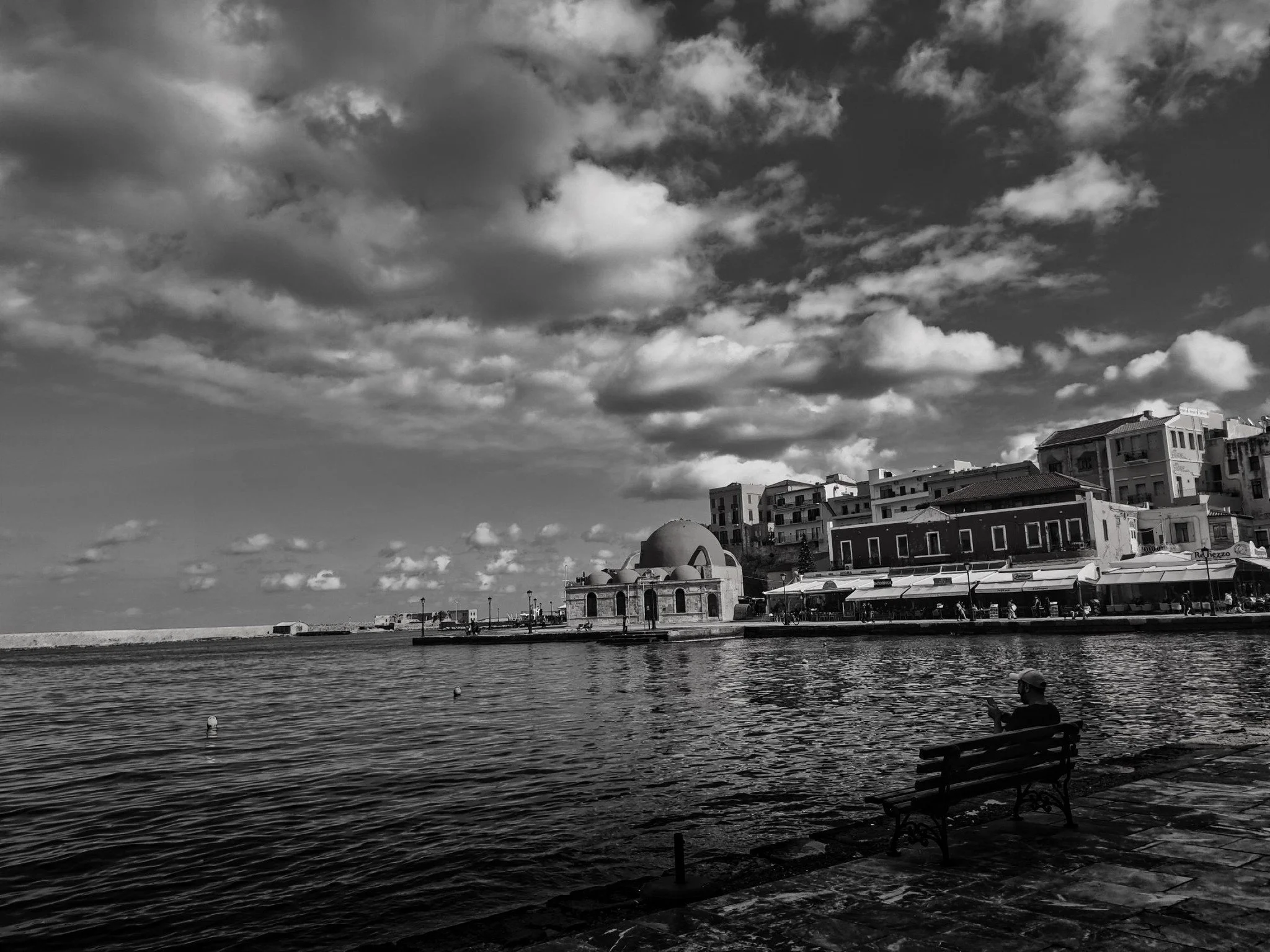 A black and white photograph of a waterfront scene with a person sitting on a bench, facing the water, with buildings and a cloudy sky in the background.