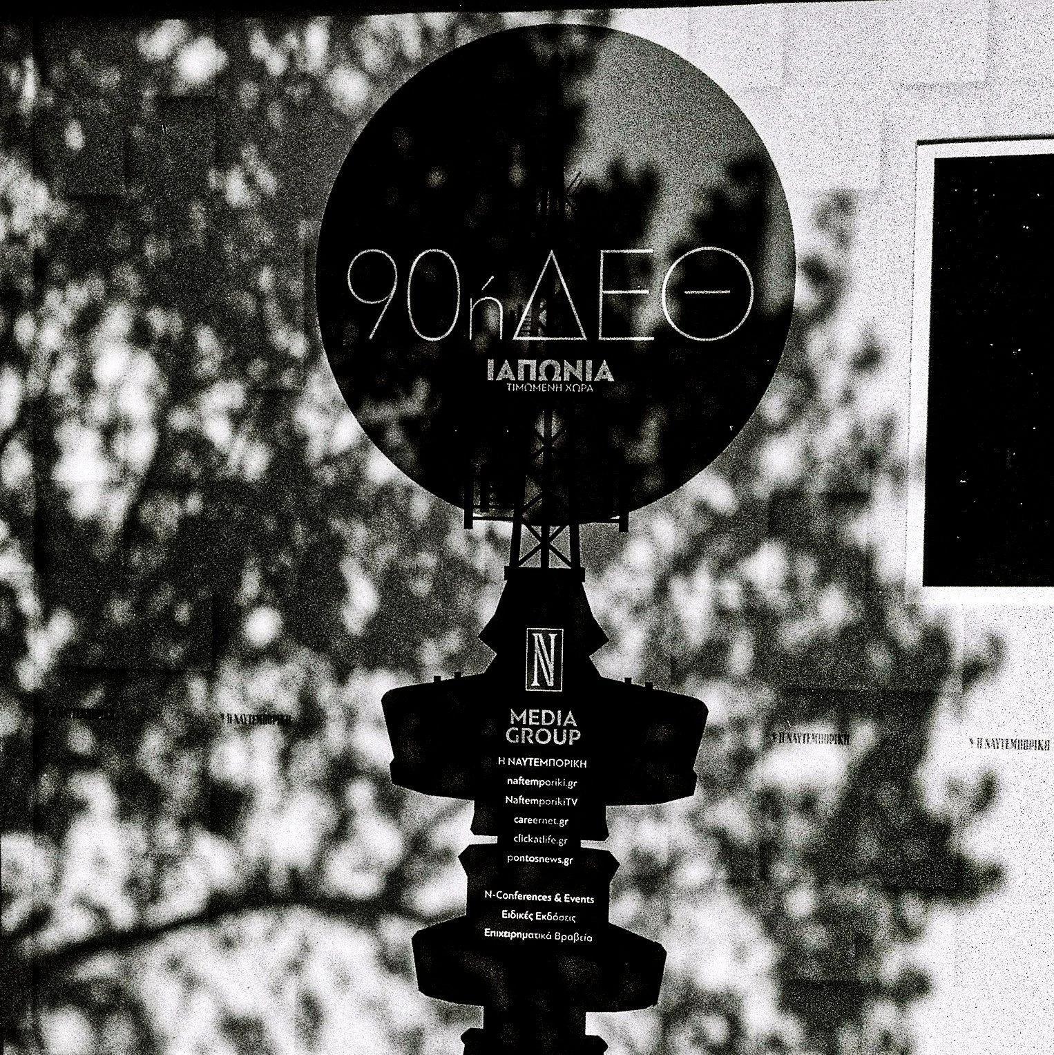 A black and white image of a weather vane with shadowed cityscape and trees in the background, displaying logos and signage for media and event companies.