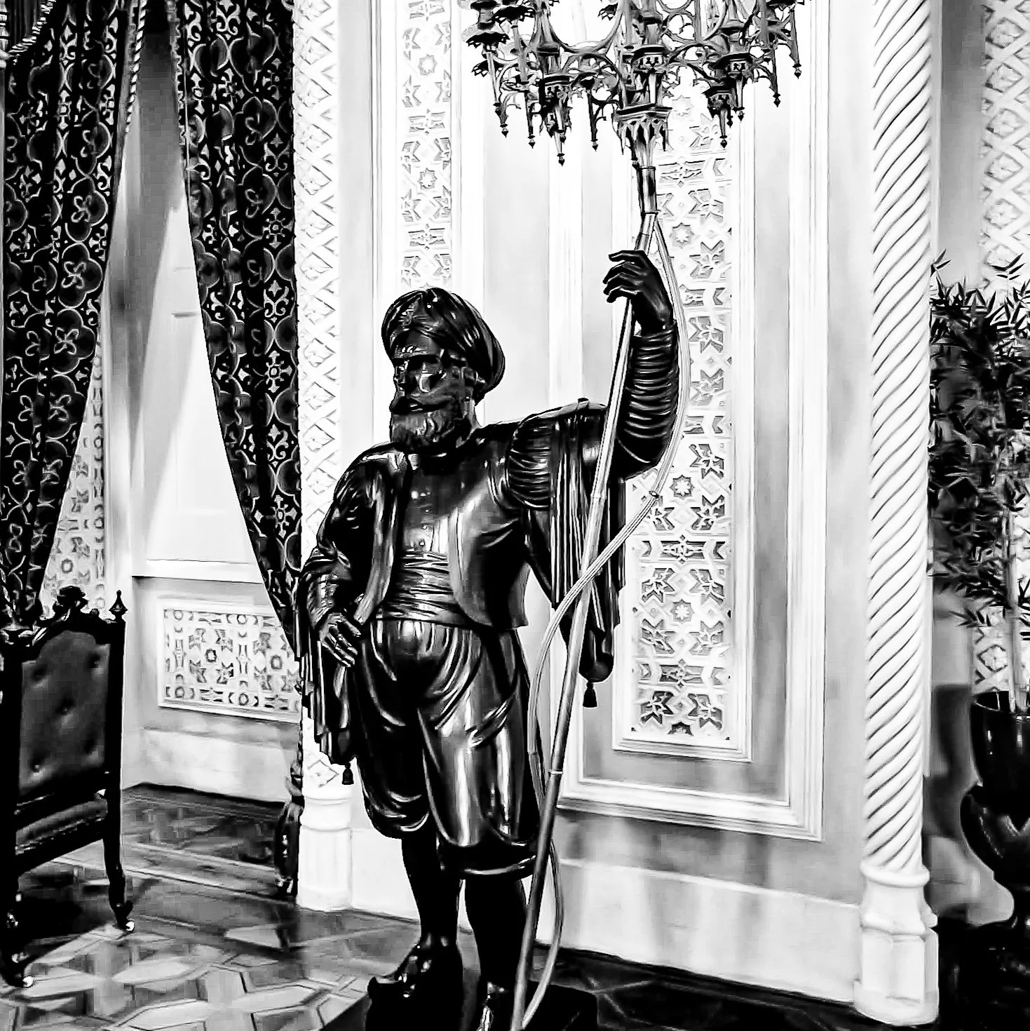 A black-and-white photo of a statue of a man in traditional Middle Eastern attire, holding a staff, inside an ornate room with patterned curtains and decorative wall panels.